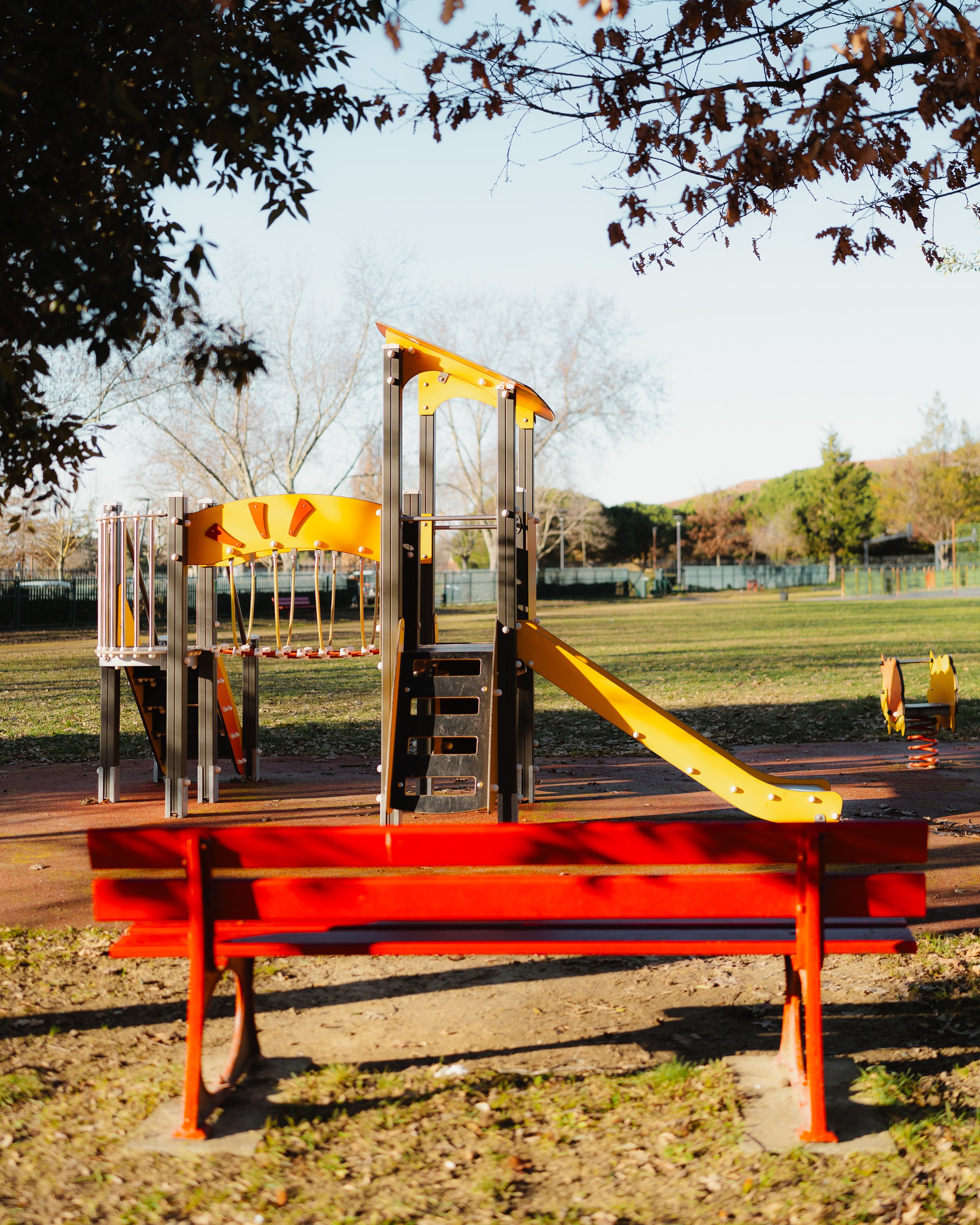 Empty playground with a red bench, yellow slide, and climbing structure in a park during daytime, with trees and a sports field in the background.