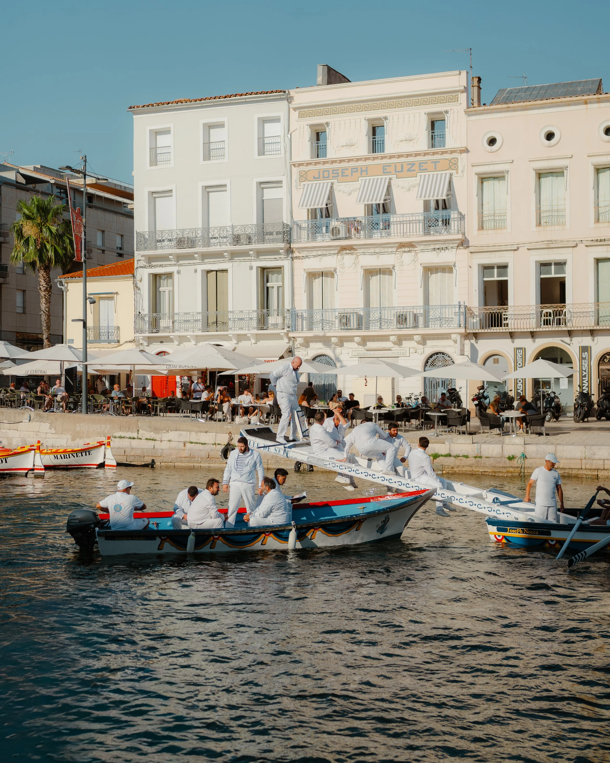 People in white clothing preparing to race boats on a river with a busy waterfront cafe and white buildings in the background.