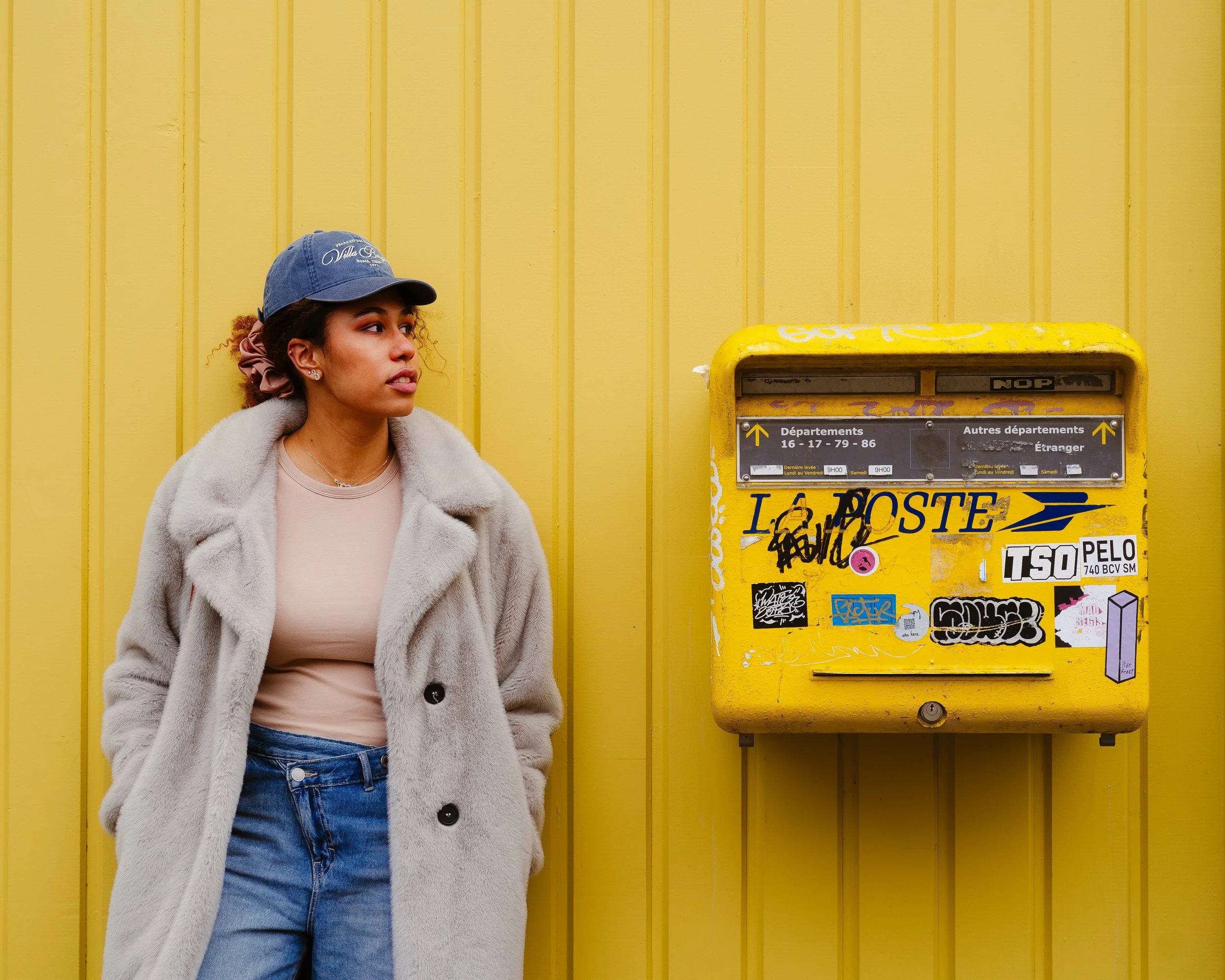 A woman wearing a beige coat, pink shirt, and blue jeans stands against a yellow wall with a yellow mailbox nearby.