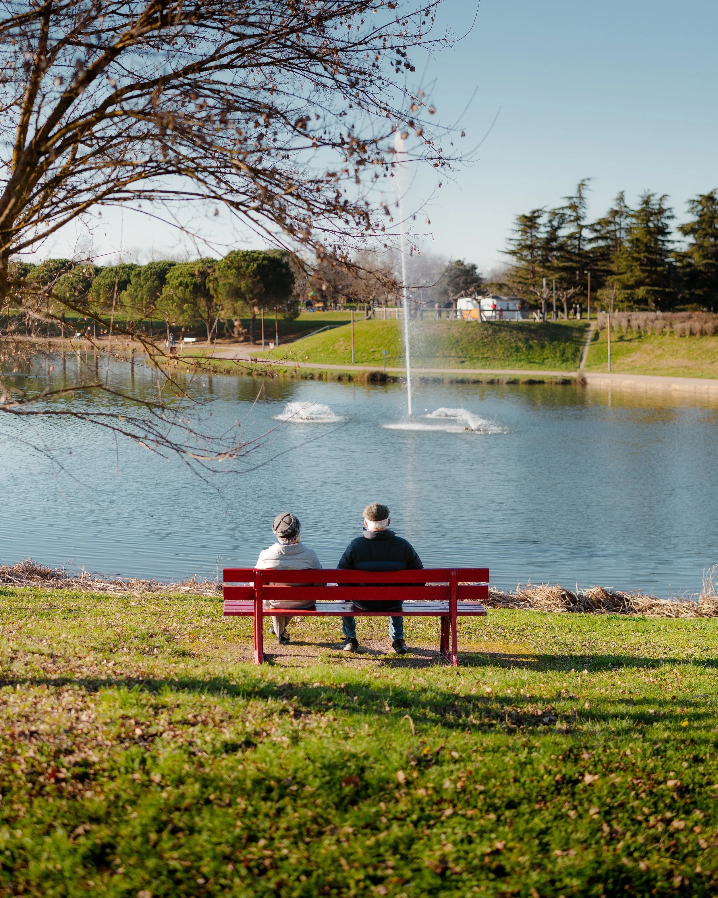 Two people sit on a red park bench watching fountains in a pond, surrounded by trees and grassy areas on a clear day.