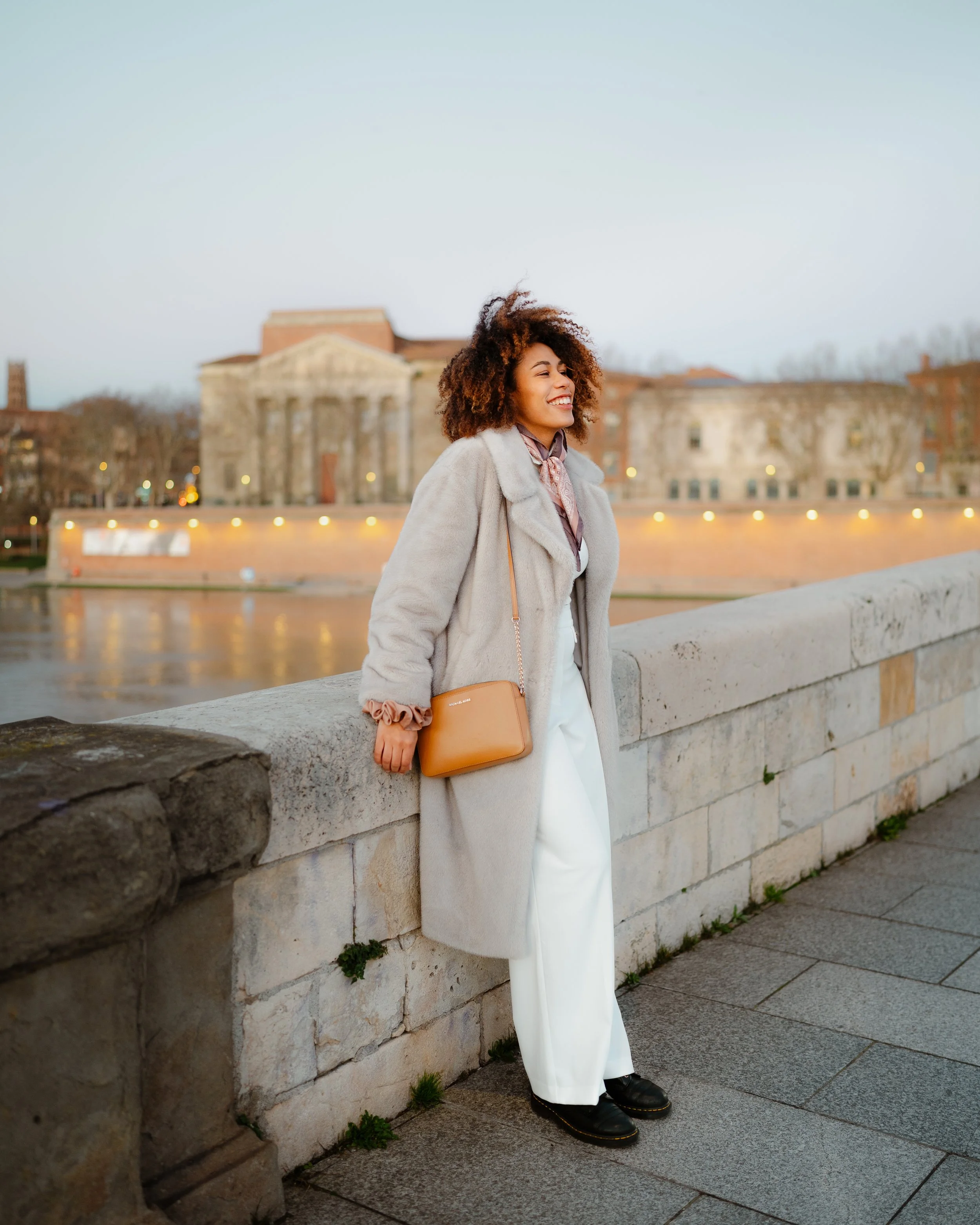 A woman with curly hair smiling and leaning against a stone railing near a river at sunset, wearing a long coat, white pants, black shoes, and carrying a small brown handbag.
