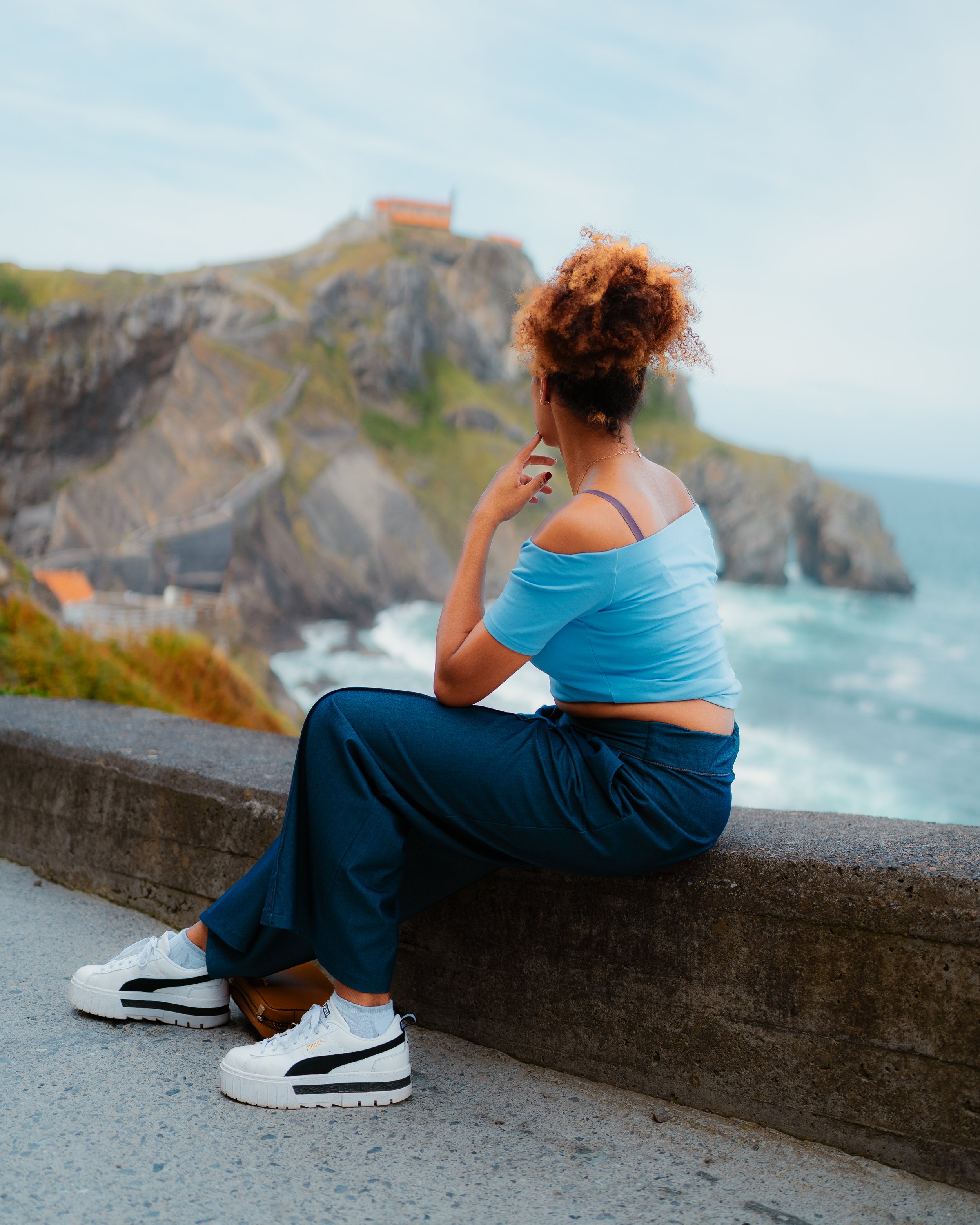 A woman sitting on a concrete ledge, looking at a coastal landscape with steep cliffs and a path leading up to a building on the top, under a clear blue sky.