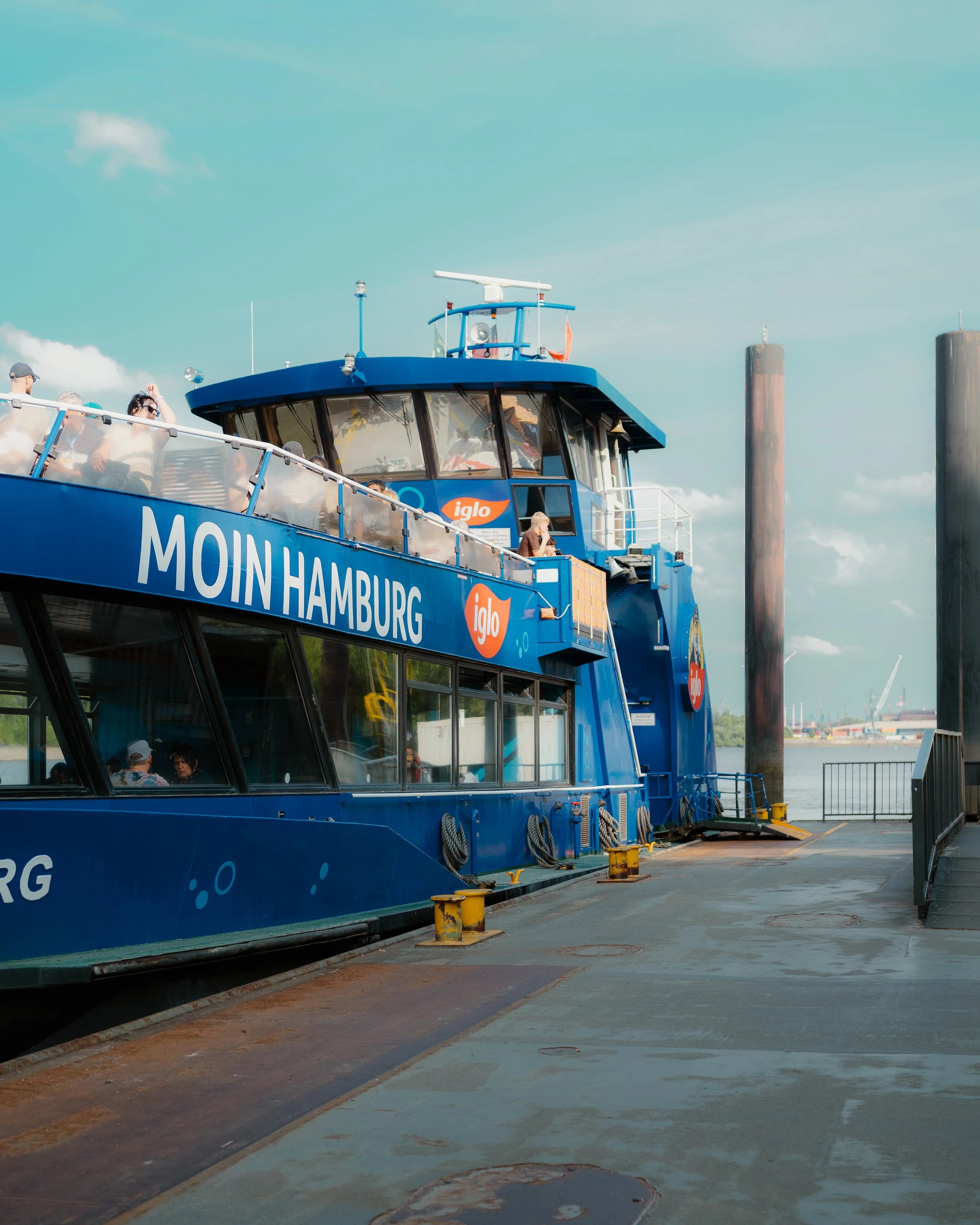 A blue ferry boat docked at the harbor with the words 'MOIN HAMBURG' on its side. Passengers are visible on the upper deck, enjoying the ride, with some taking photos. The sky is partly cloudy, and there are large industrial posts in the background.