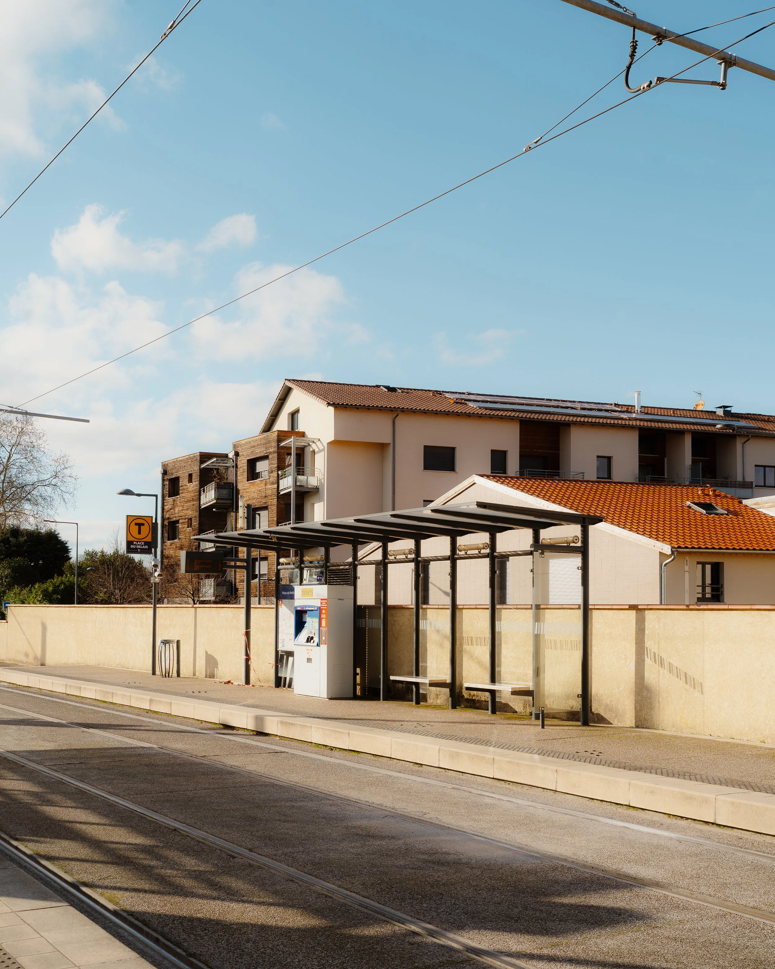 A tram stop on a city street with a covered waiting area, vending machines, and a beige fence. Residential buildings with tiled roofs are in the background under a blue sky with clouds.