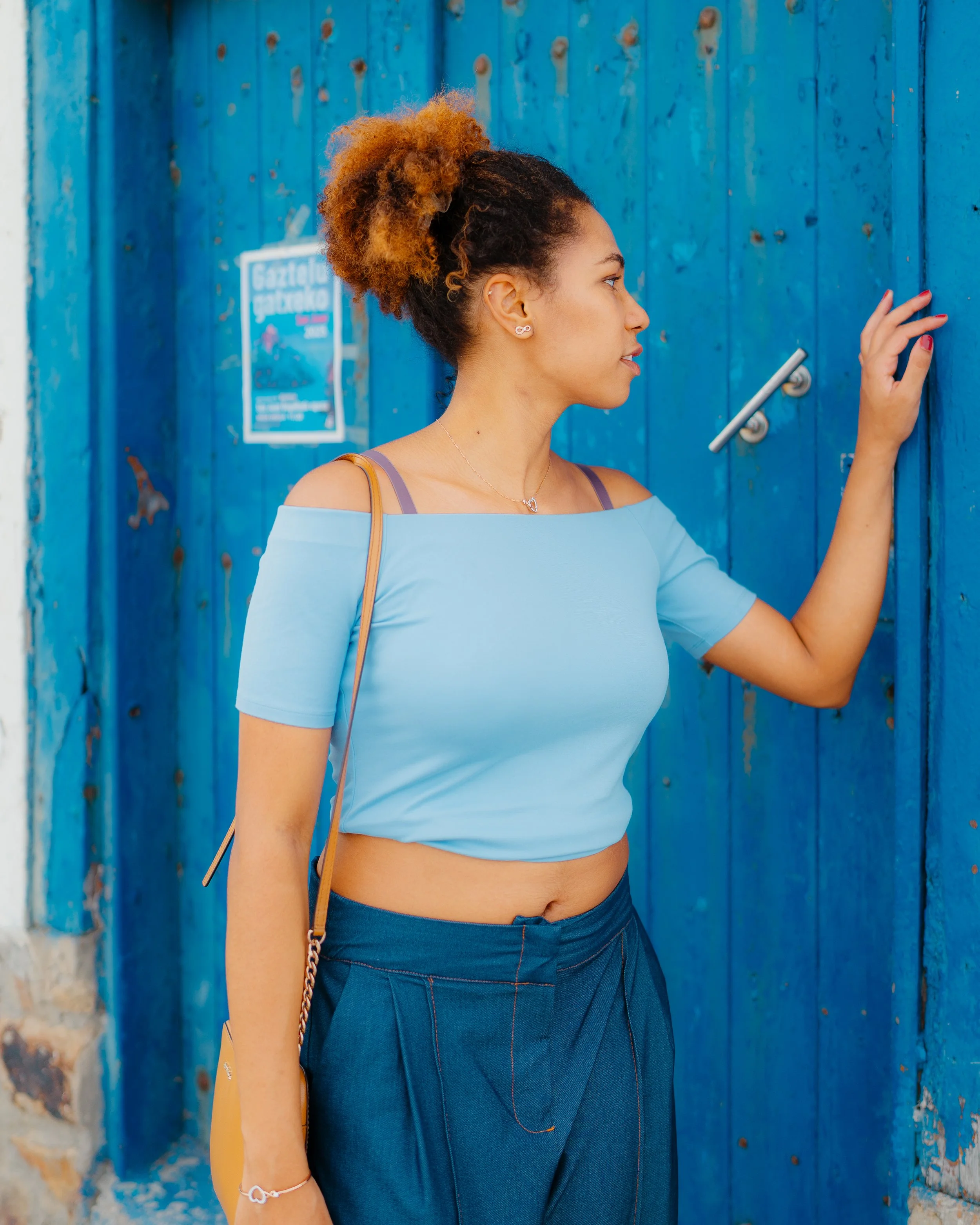 Young woman with natural hair touching a blue wooden door, wearing a light blue off-shoulder top and dark blue pants.