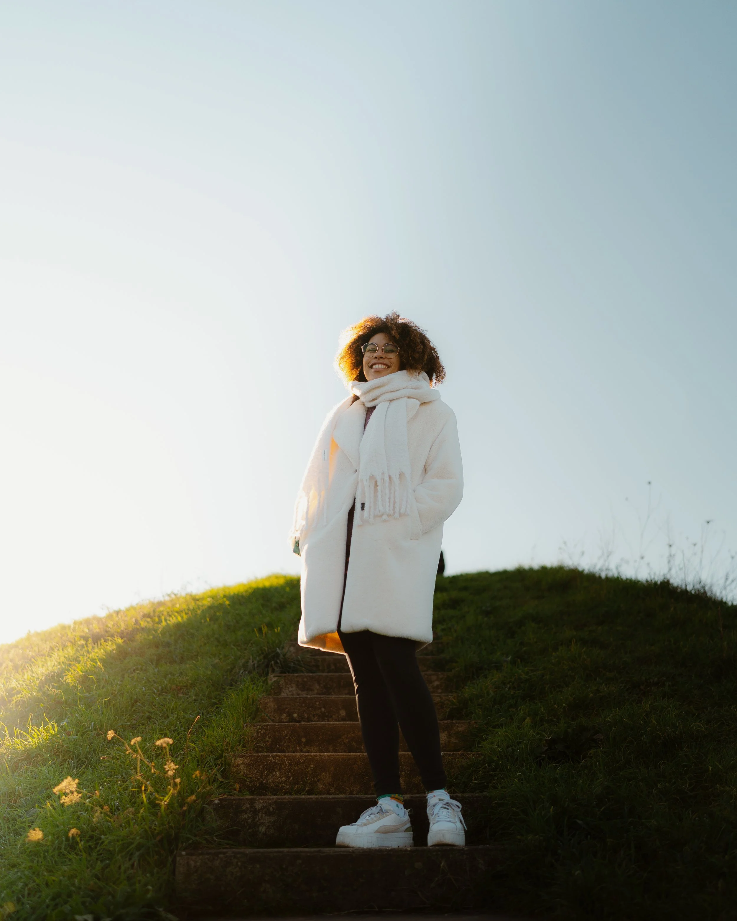 A woman wearing glasses and a white coat standing on outdoor stairs, smiling with sunlight behind her.