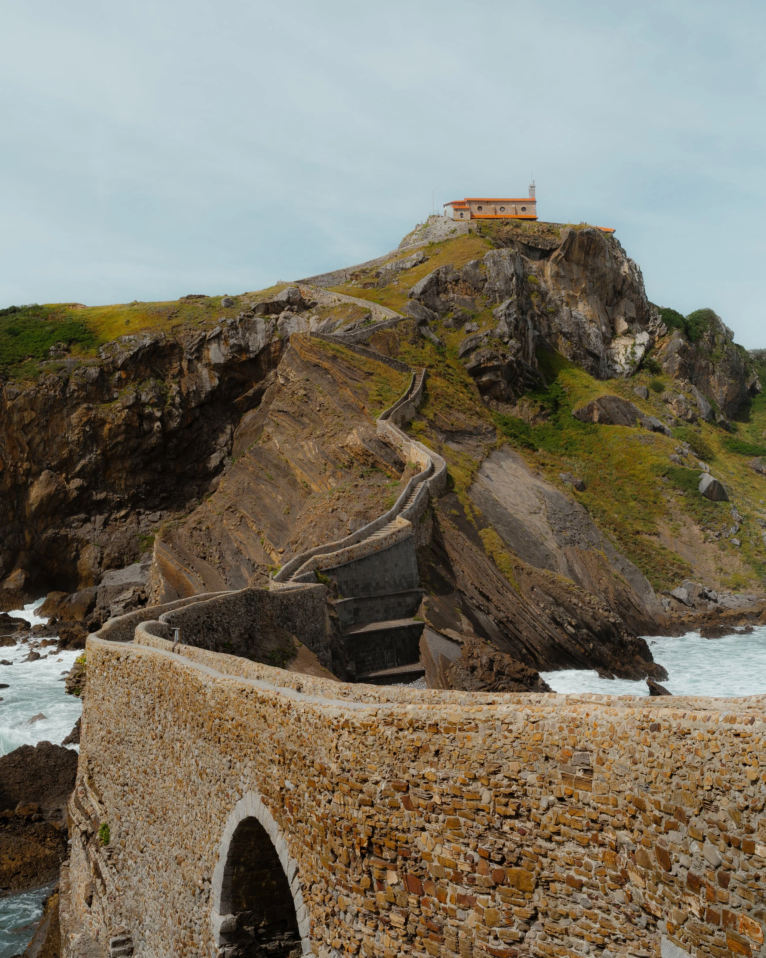 A winding stone staircase leading up a rocky cliff to a building at the top, with a stone arch bridge near the ocean in the foreground.