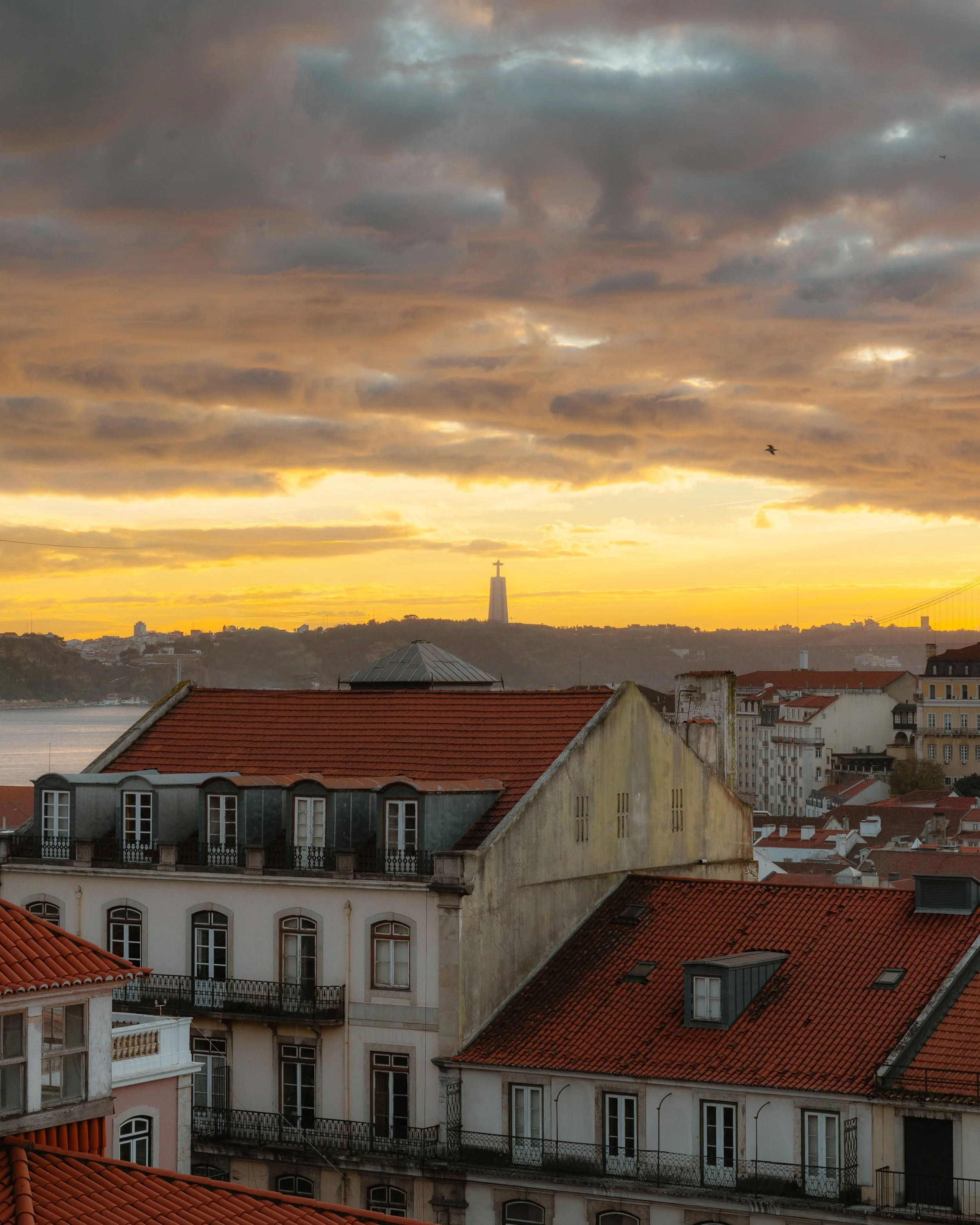 Sunset over Lisbon with clouds, rooftops with red tiles, and the Christ the King monument in the distance.