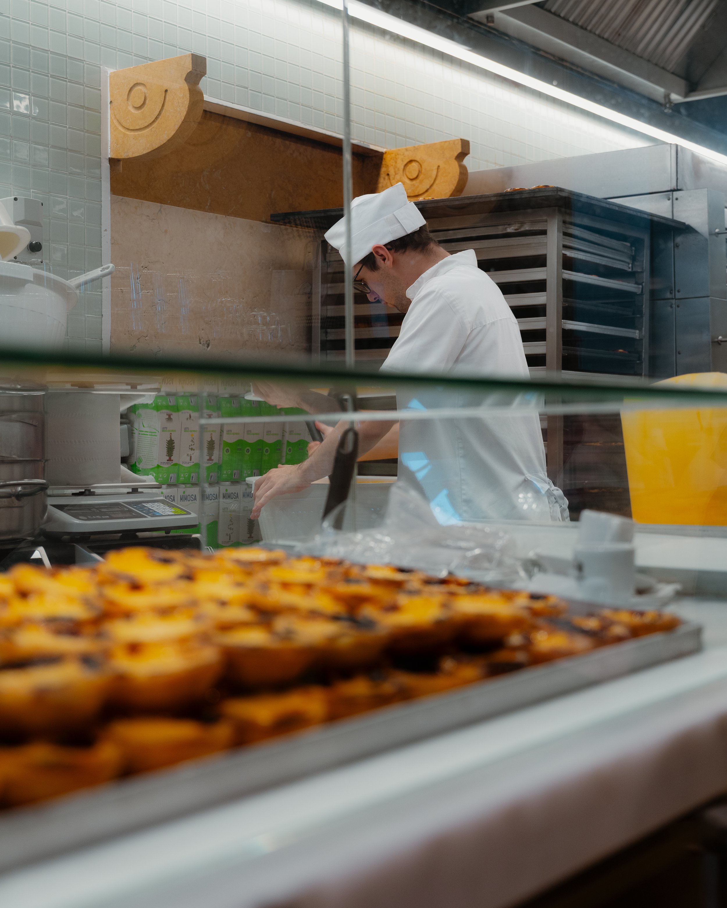 A chef in a white uniform and hat working behind a glass display case, with baked pastries in the foreground.