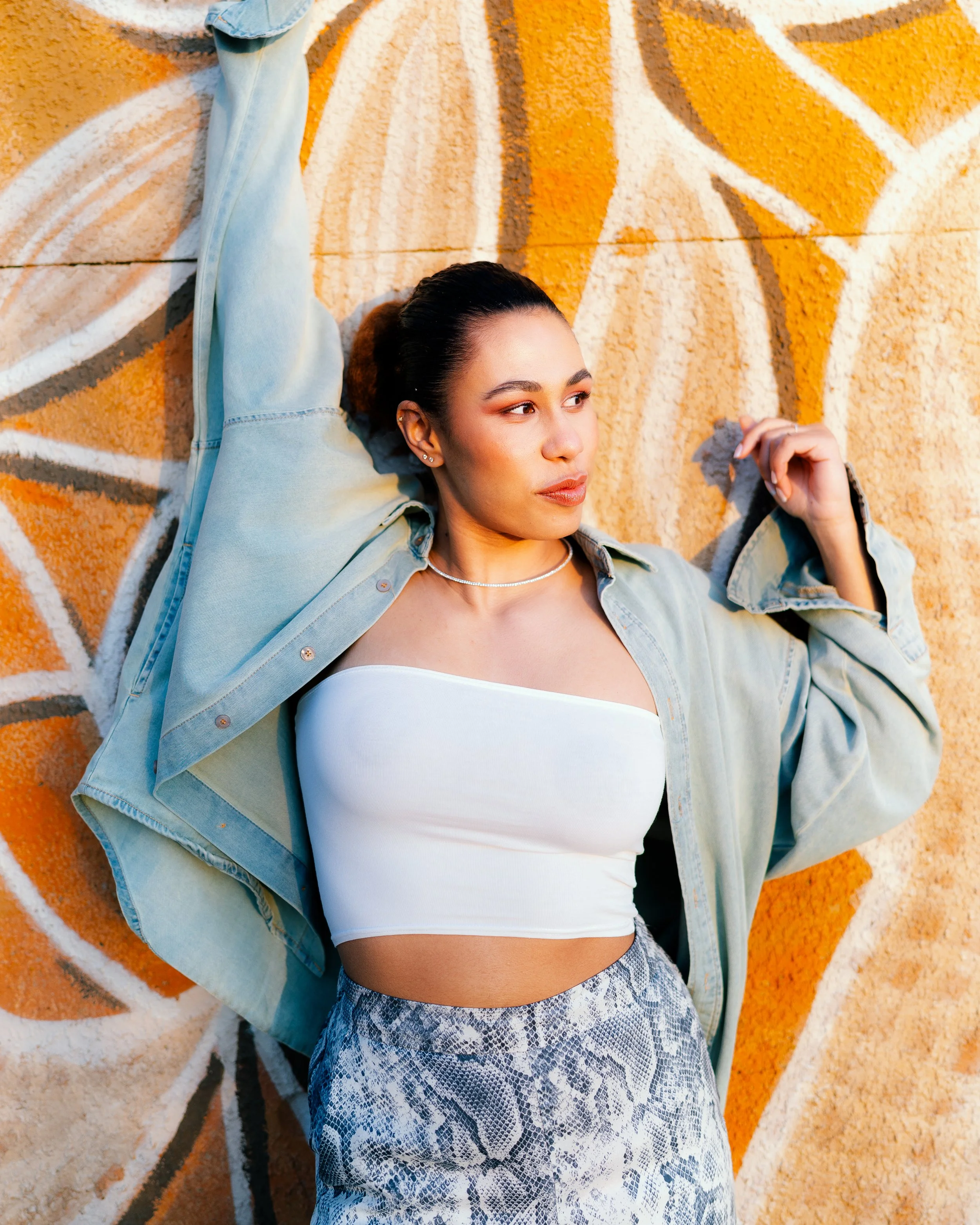 A young woman with dark hair tied back, wearing a white strapless top, a light denim jacket, and snakeskin patterned pants, leaning against a colorful graffiti wall.