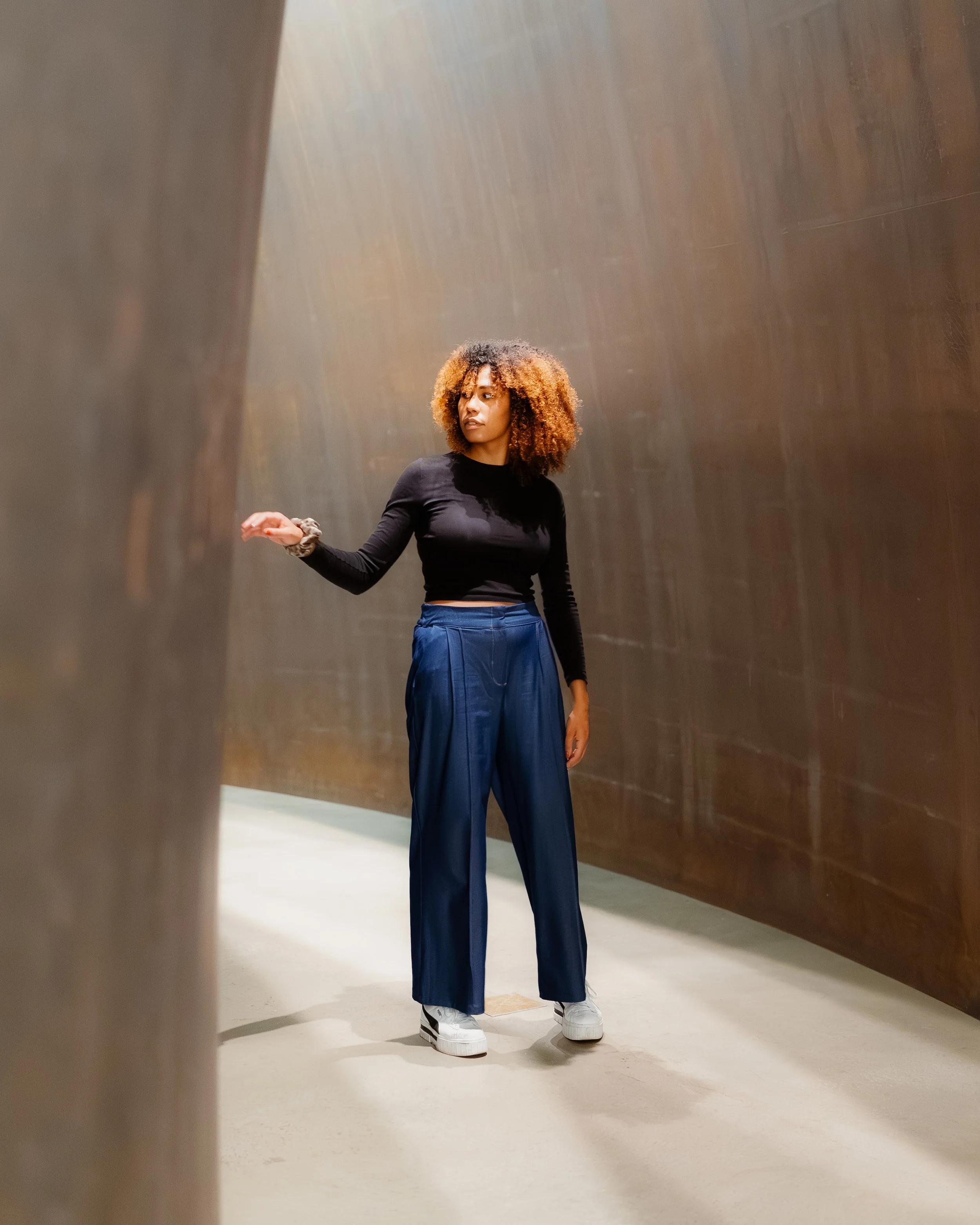 Young woman with curly hair wearing a black long-sleeve shirt, blue pants, and white shoes standing in a narrow, curved metallic corridor.