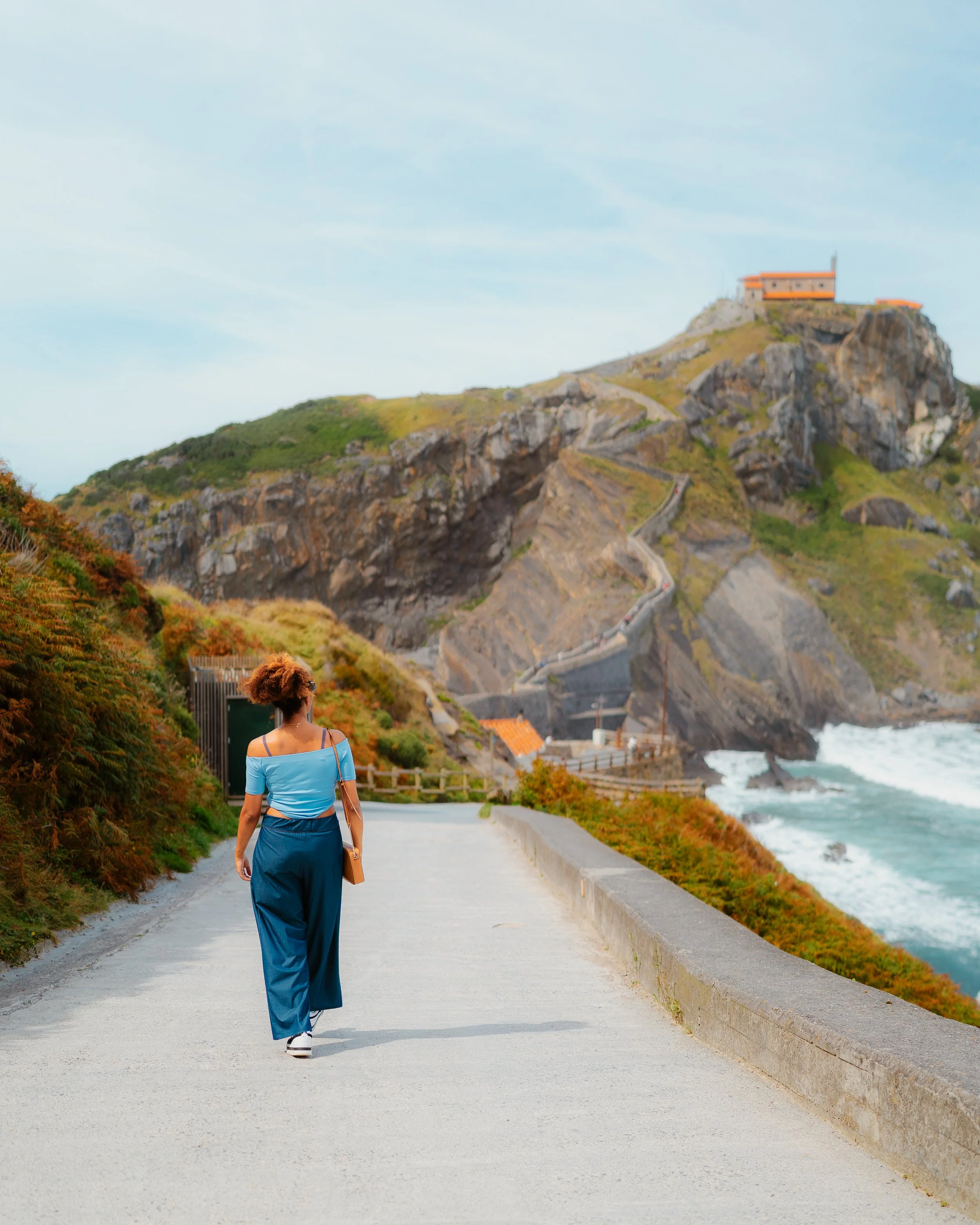 A woman dressed in a blue off-shoulder top and wide-leg pants walks on a coastal pathway with cliffs, a winding staircase, and a house on a hill in the background.