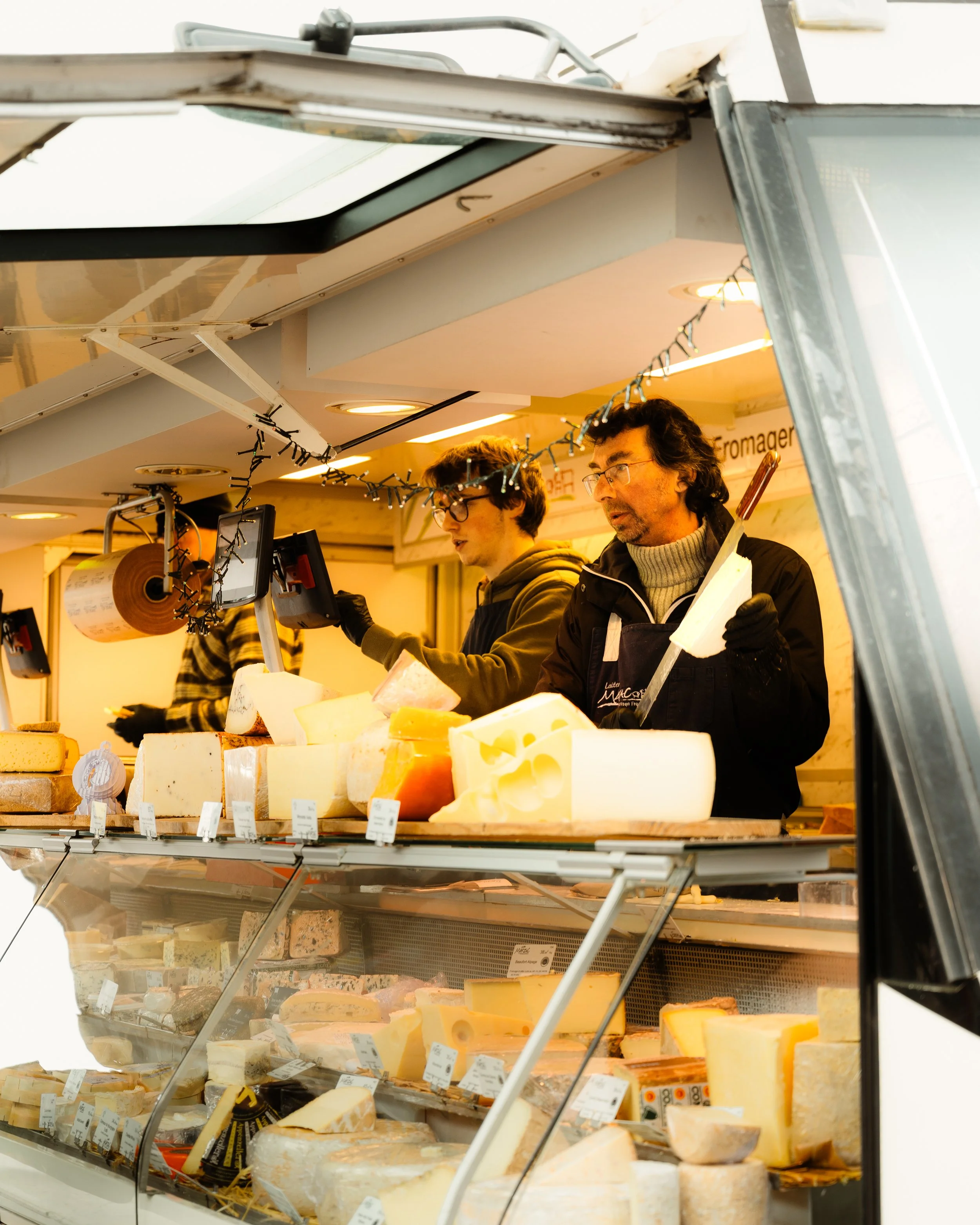Two men work behind a cheese display counter at a cheese shop or market, slicing and preparing cheese for sale.