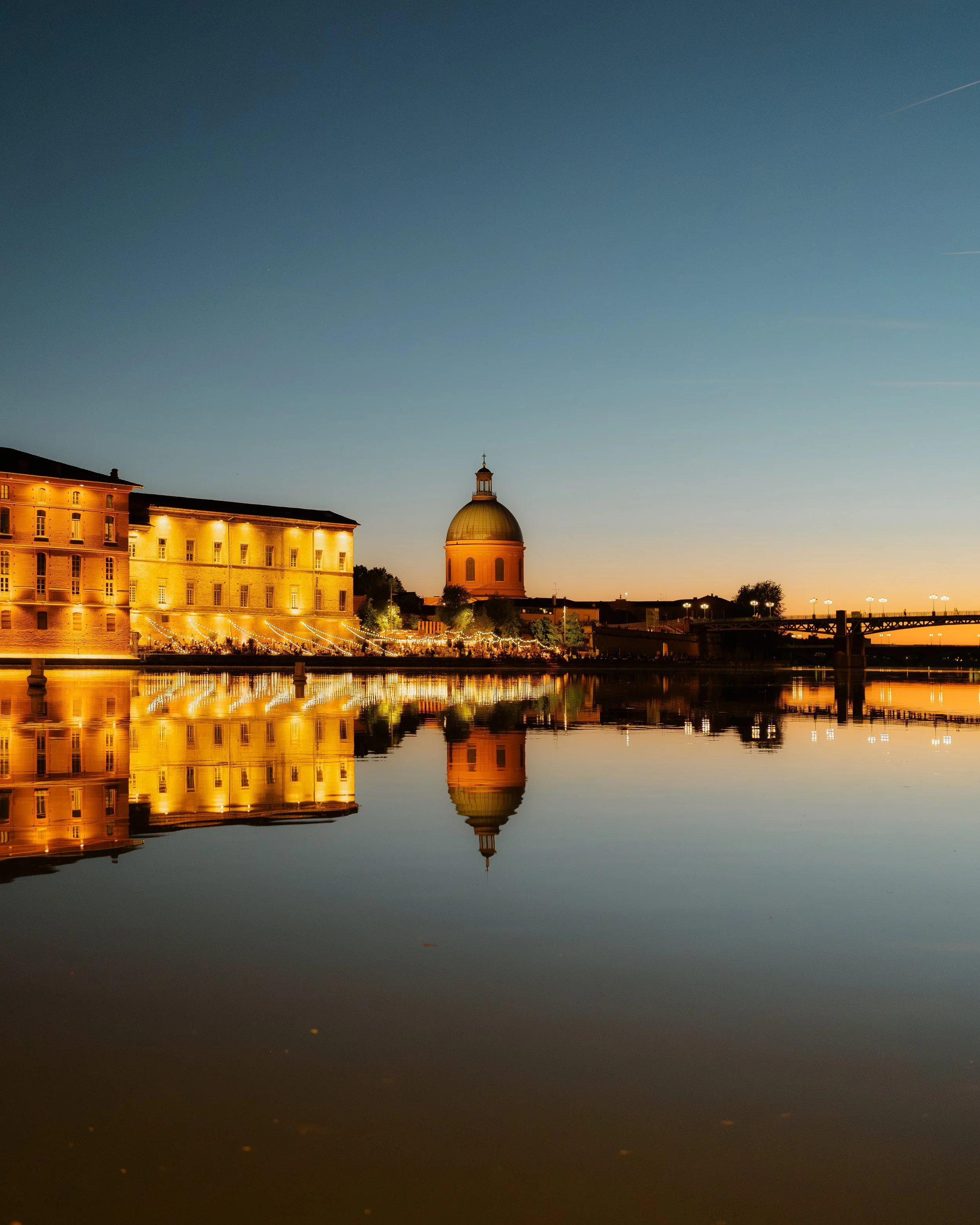 The image shows a cityscape at dusk with a historic building featuring a dome and a smaller tower reflected in calm water, with the sky darkening