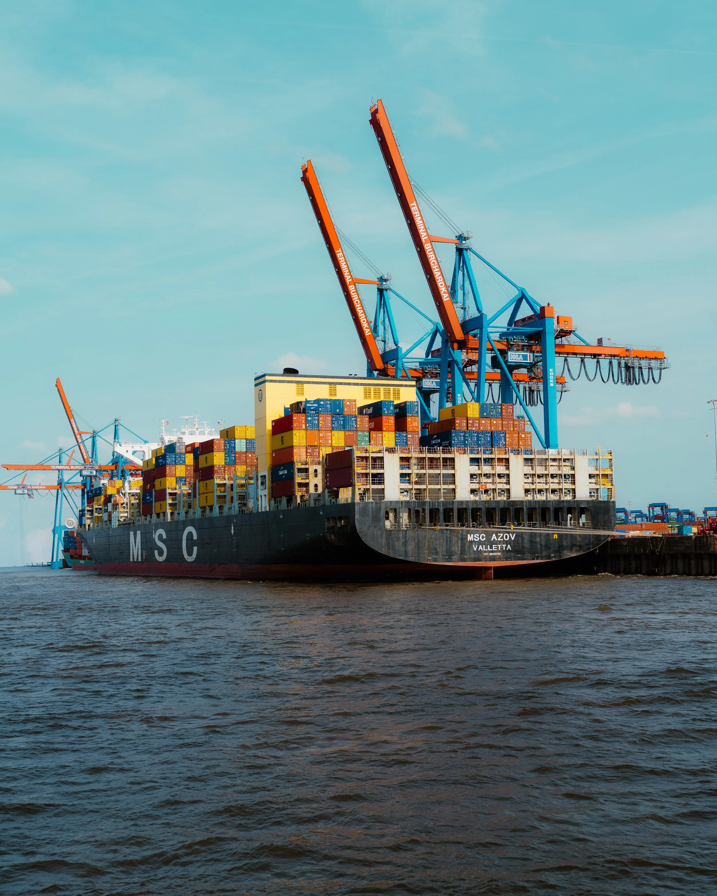 A cargo ship loaded with colorful containers docked at a port with blue cranes used for loading and unloading shipping containers.