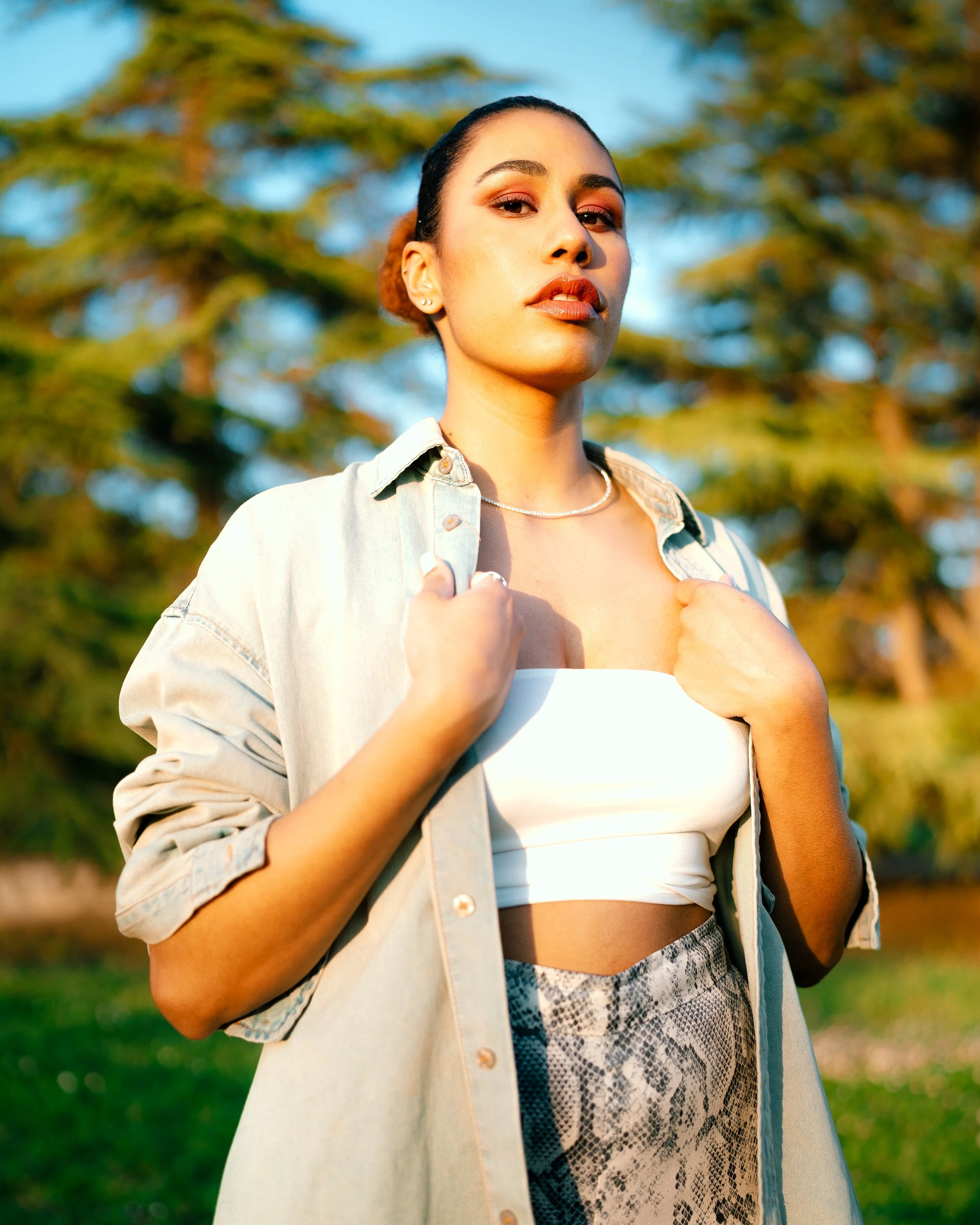 A young woman outdoors with trees and blue sky in the background, wearing a white top, a light denim shirt, and snake print pants, holding the collar of her shirt.