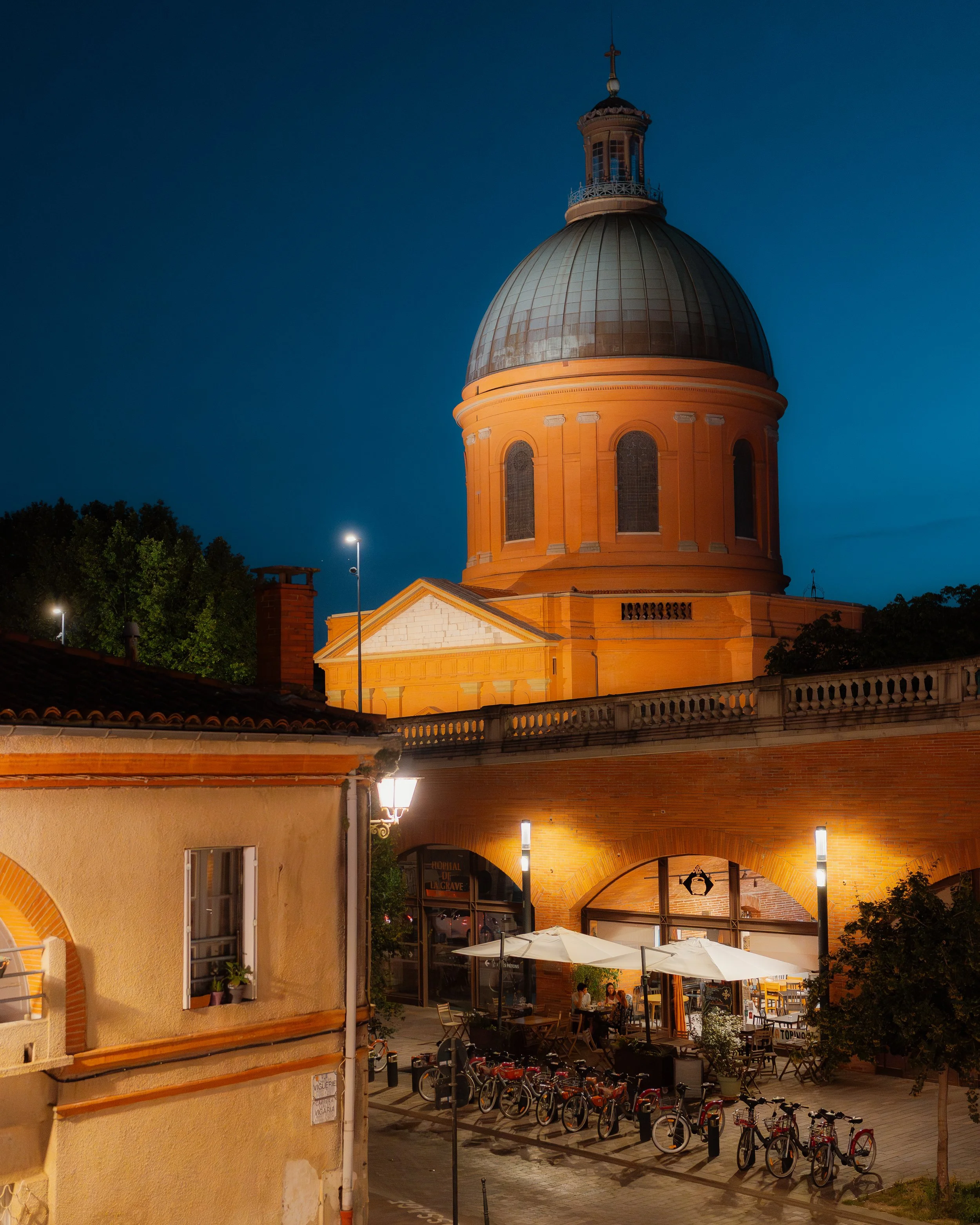 A large church with a prominent dome illuminated in warm yellow light, at night. Below the church, there is an outdoor seating area with tables, umbrellas, and people dining. Bicycles are parked near the seating area, and the scene is lit by street l