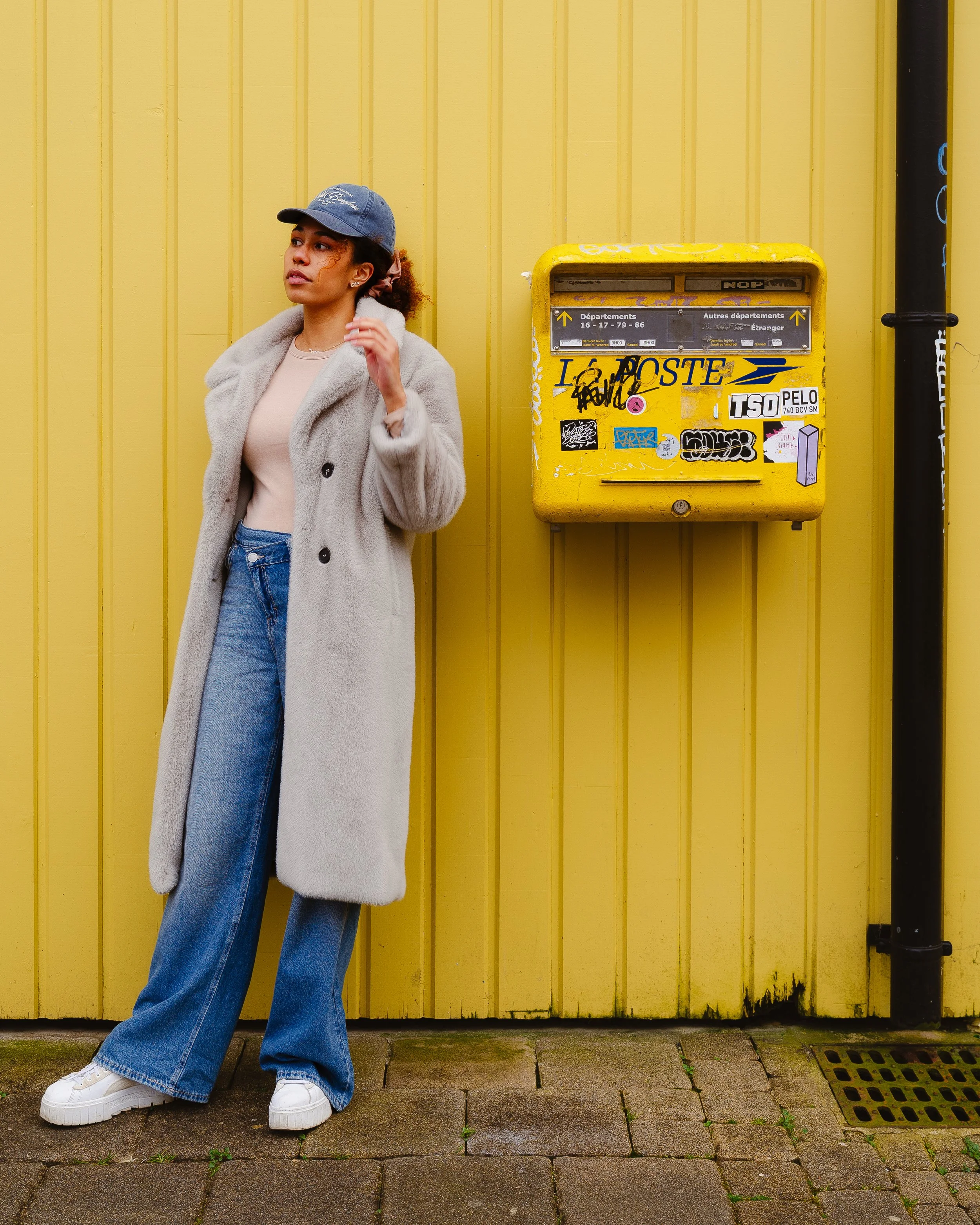 A woman standing against a yellow wall next to a yellow mailbox covered in stickers.