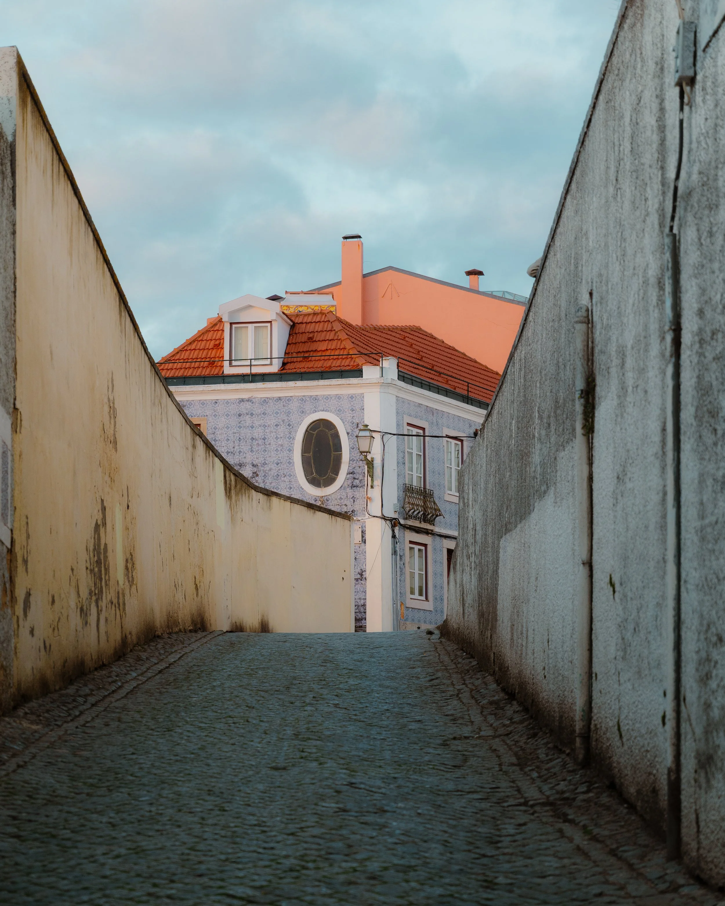A narrow, cobblestone alleyway between aged, off-white and gray walls, leading to a colorful building with a red-tiled roof and rounded window, under a partly cloudy sky.