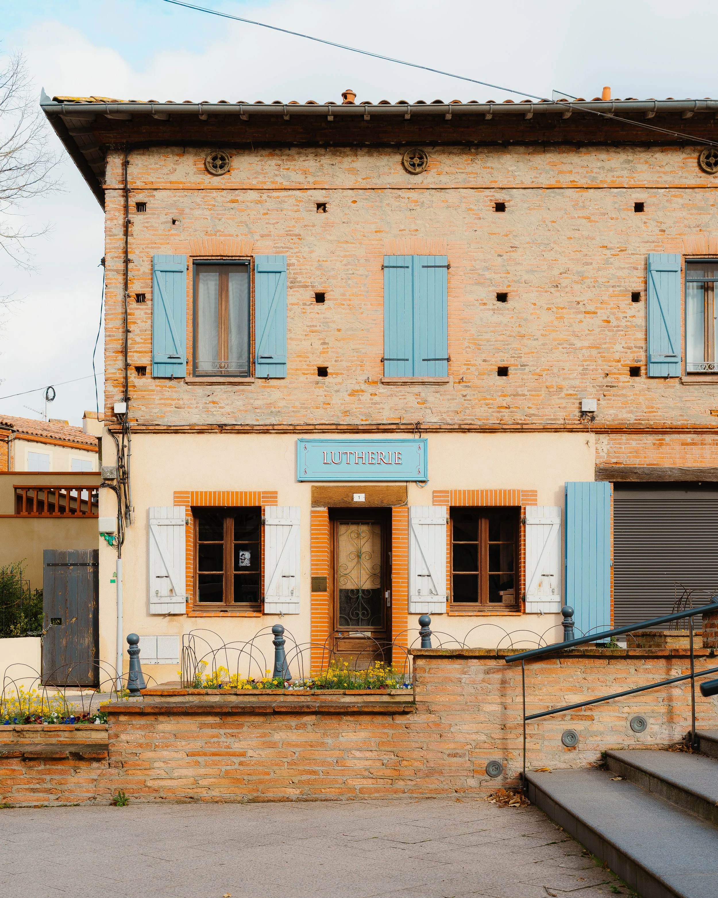 A two-story brick house with blue shutters on the windows, a sign that reads 'LUTHERIE,' and a small flower garden in front, with steps and a railing to the entrance.