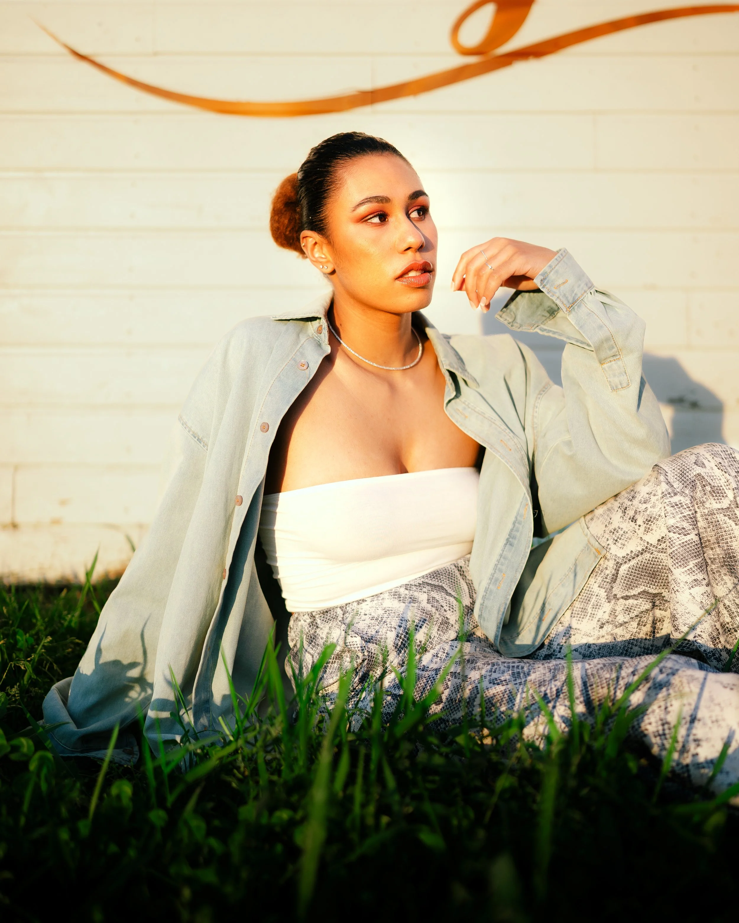 A young woman with dark hair in a bun, wearing a strapless top, a light denim jacket, and snake print pants, sitting on grass in front of a white wooden wall with a metal decorative element, bathed in warm sunlight.