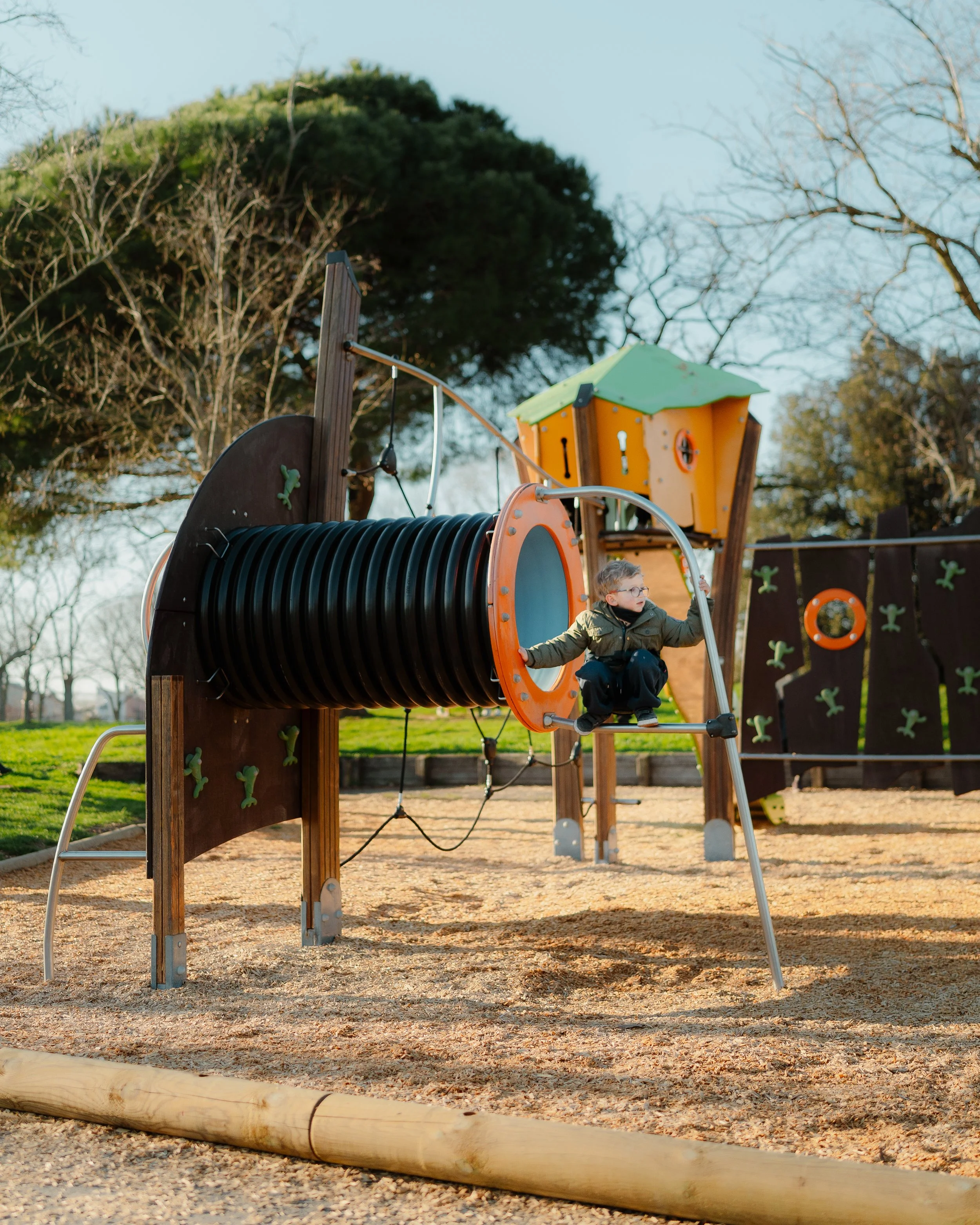A young boy on a playground climbing through a plastic tunnel attached to a wooden play structure with trees in the background.