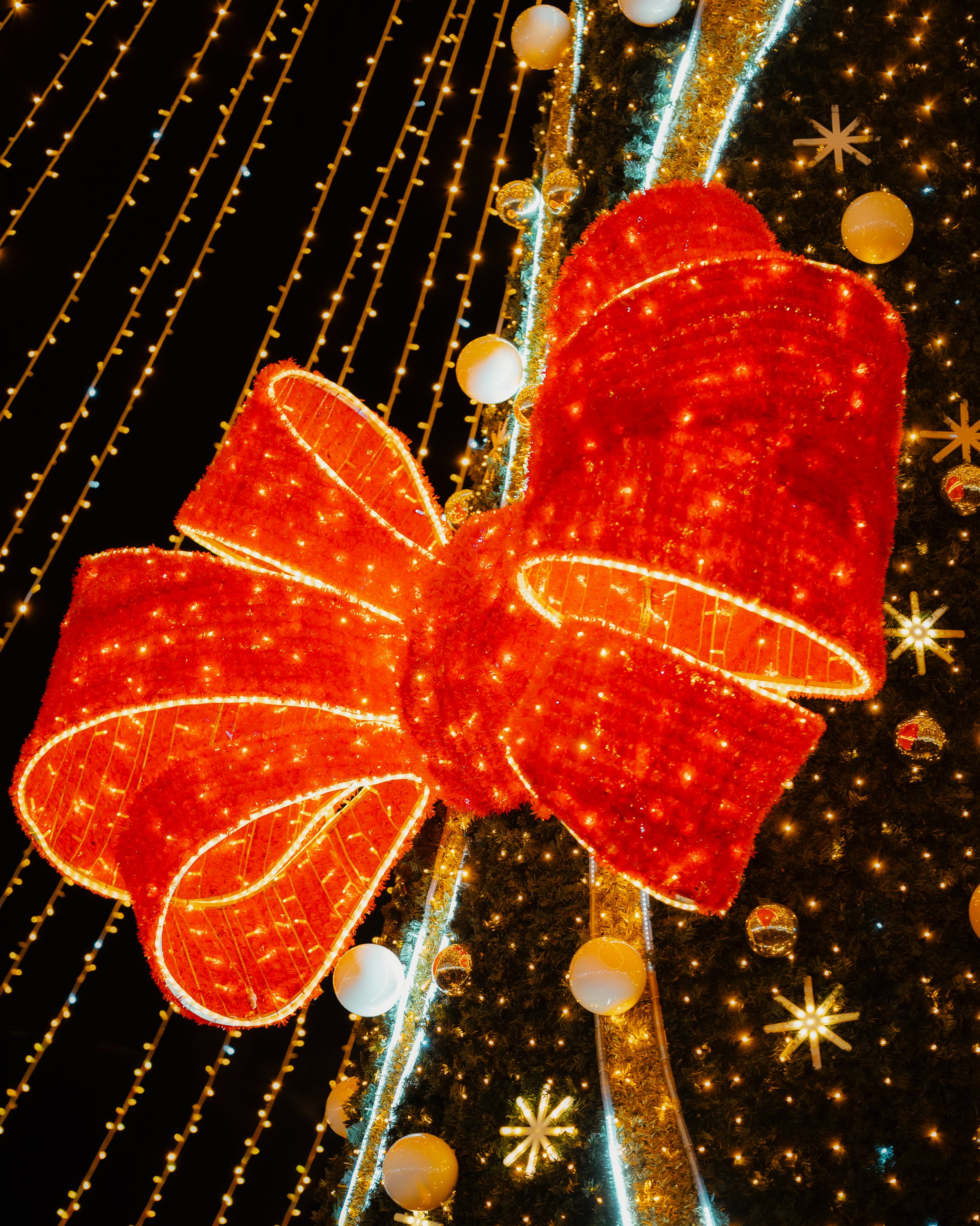 Large red illuminated bow decoration on a Christmas tree with gold and white ornaments, set against a backdrop of yellow string lights and star-shaped lights.
