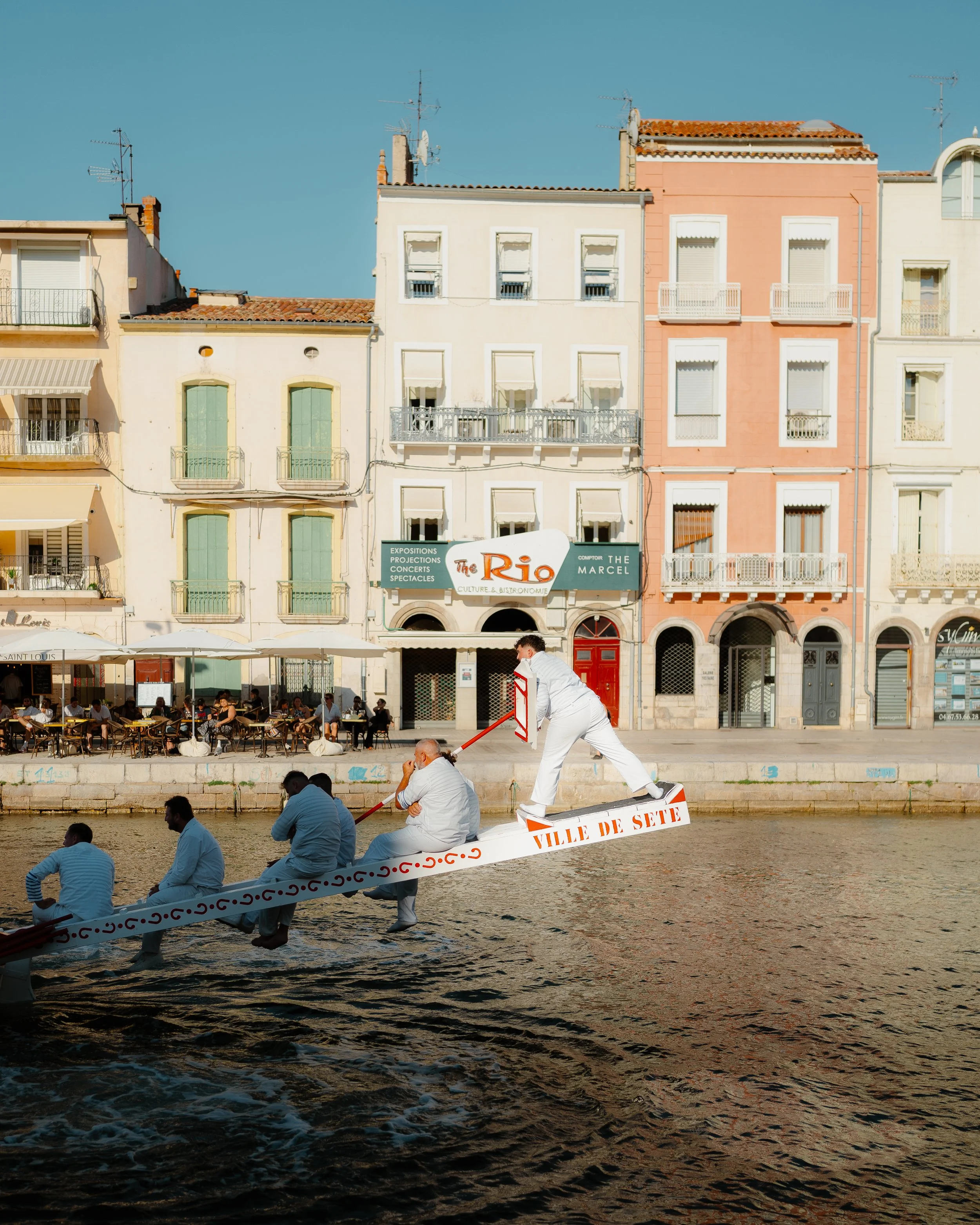 A group of people in white clothes on a boat, with one person standing and holding a paddle, in front of a row of colorful buildings and a restaurant with outdoor seating by the water.