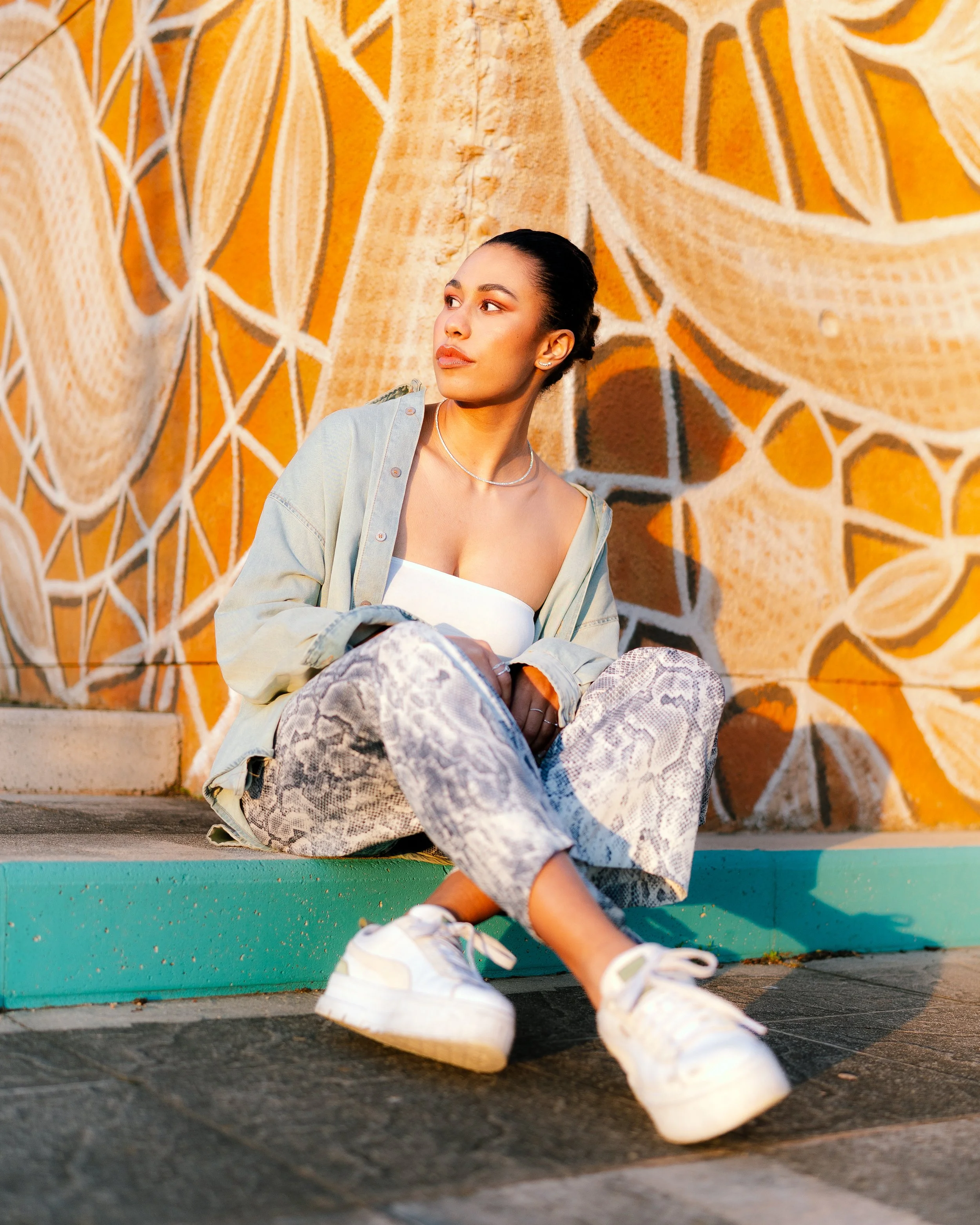 A young woman with dark hair tied back, sitting on the ground against a colorful abstract mural, wearing a white top, light jacket, snake-print pants, and white sneakers, looking off to the side.