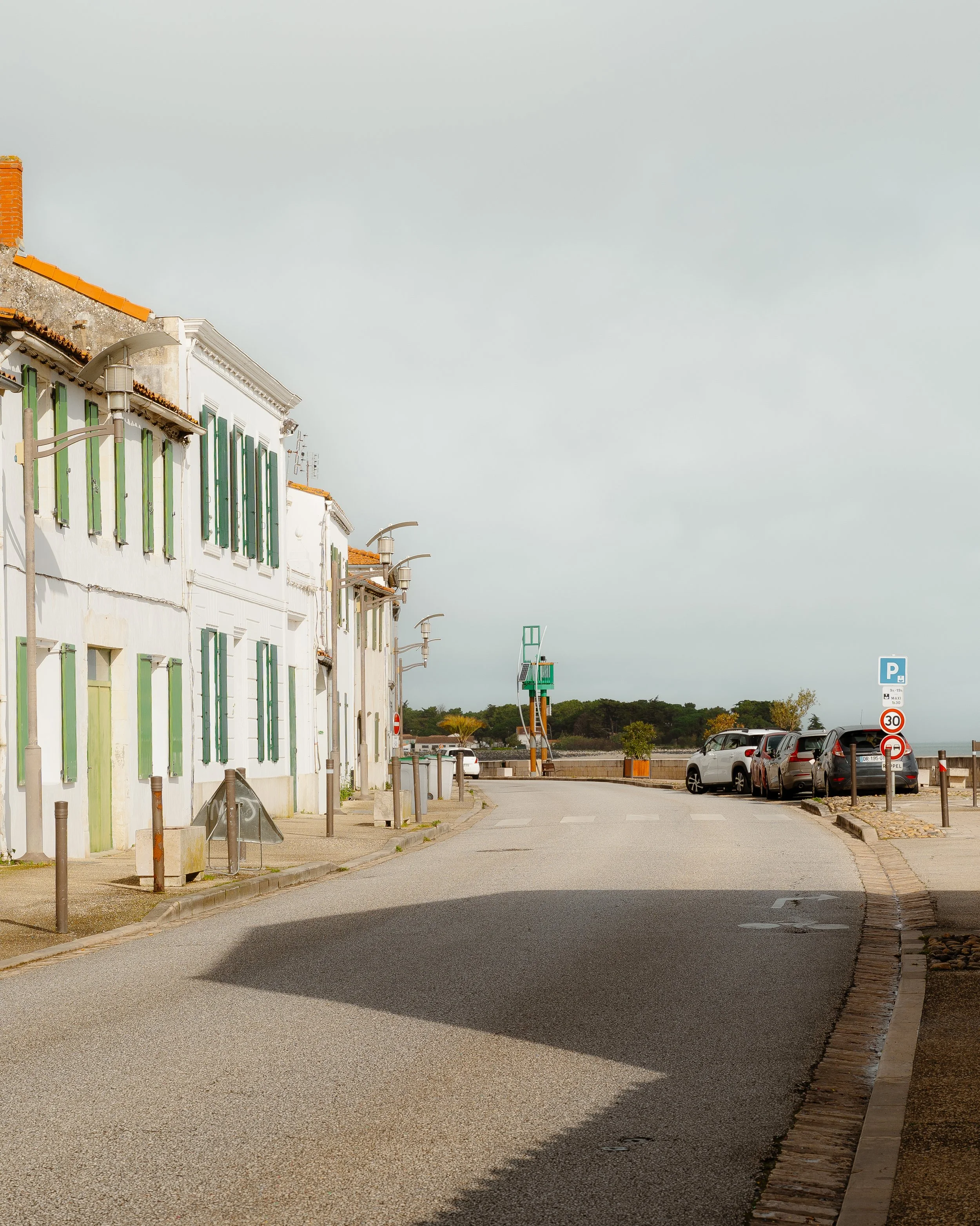A quiet street with parked cars near the coast, old buildings with green and white shutters, parking signs, and a cloudy sky.