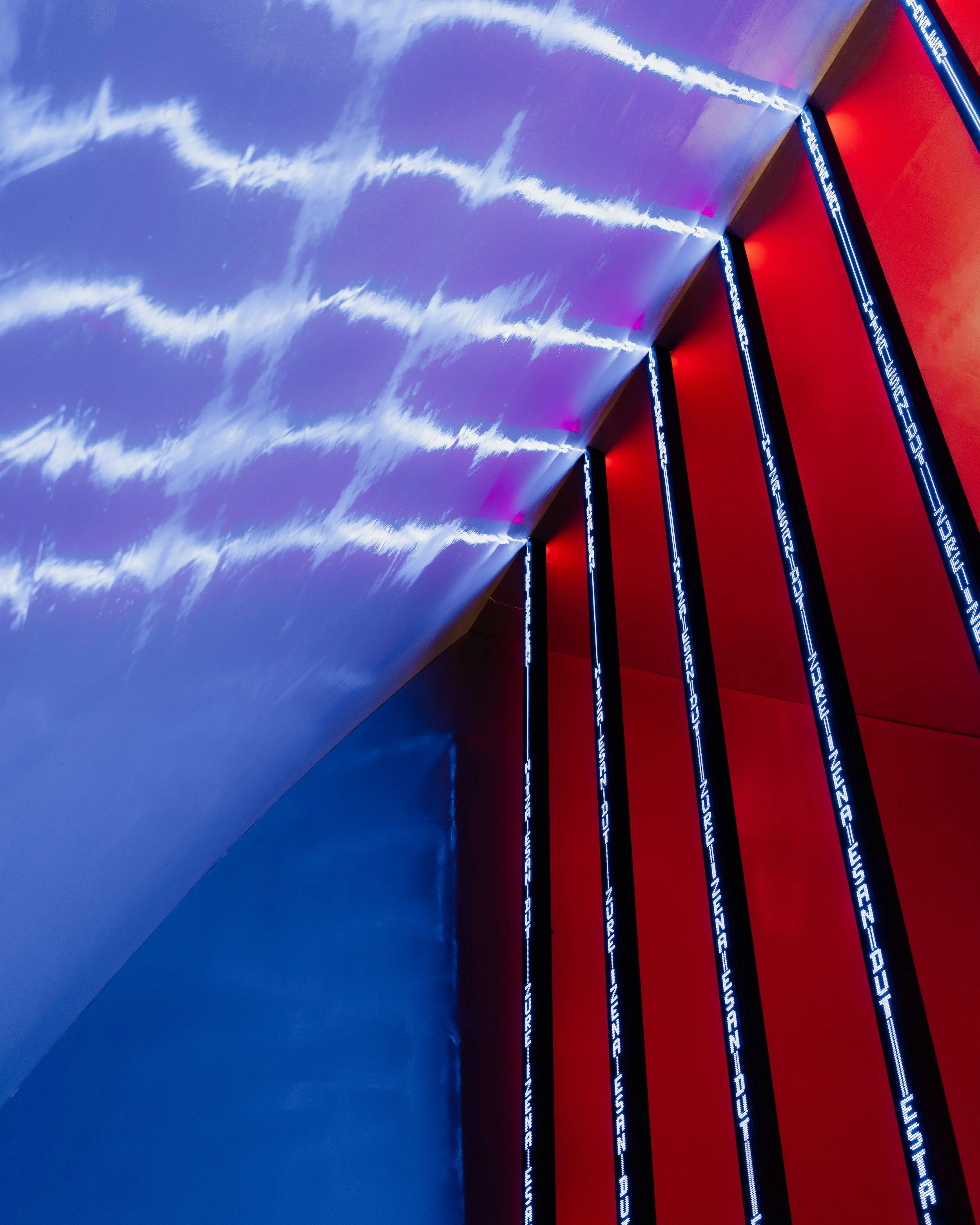 Interior art installation with blue LED lightbars on red background, displaying digital text and lightning images projected on ceiling.