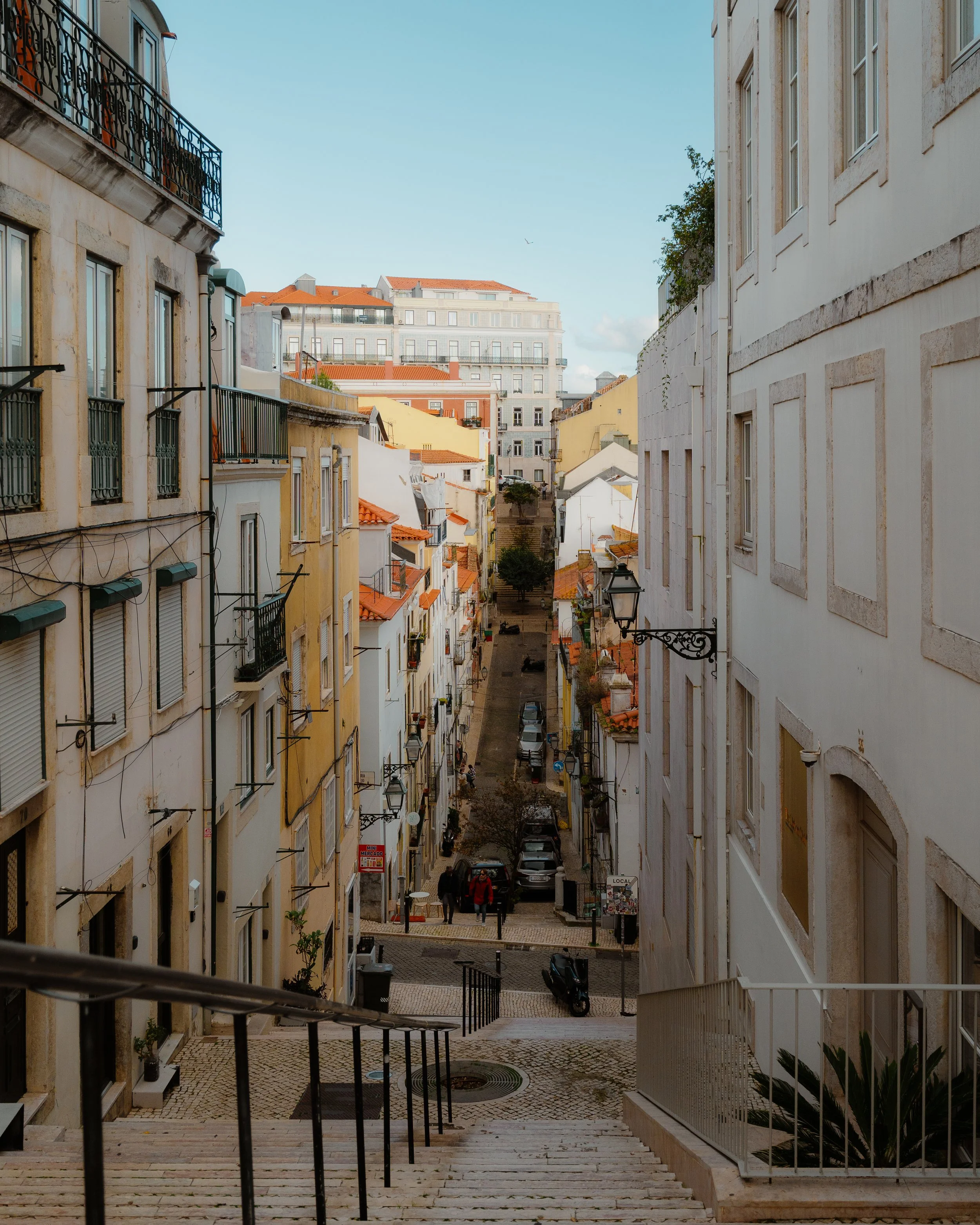 Photograph of a narrow city street viewed from top of a staircase, with apartment buildings on both sides, parked cars, and a few pedestrians, under a clear blue sky.