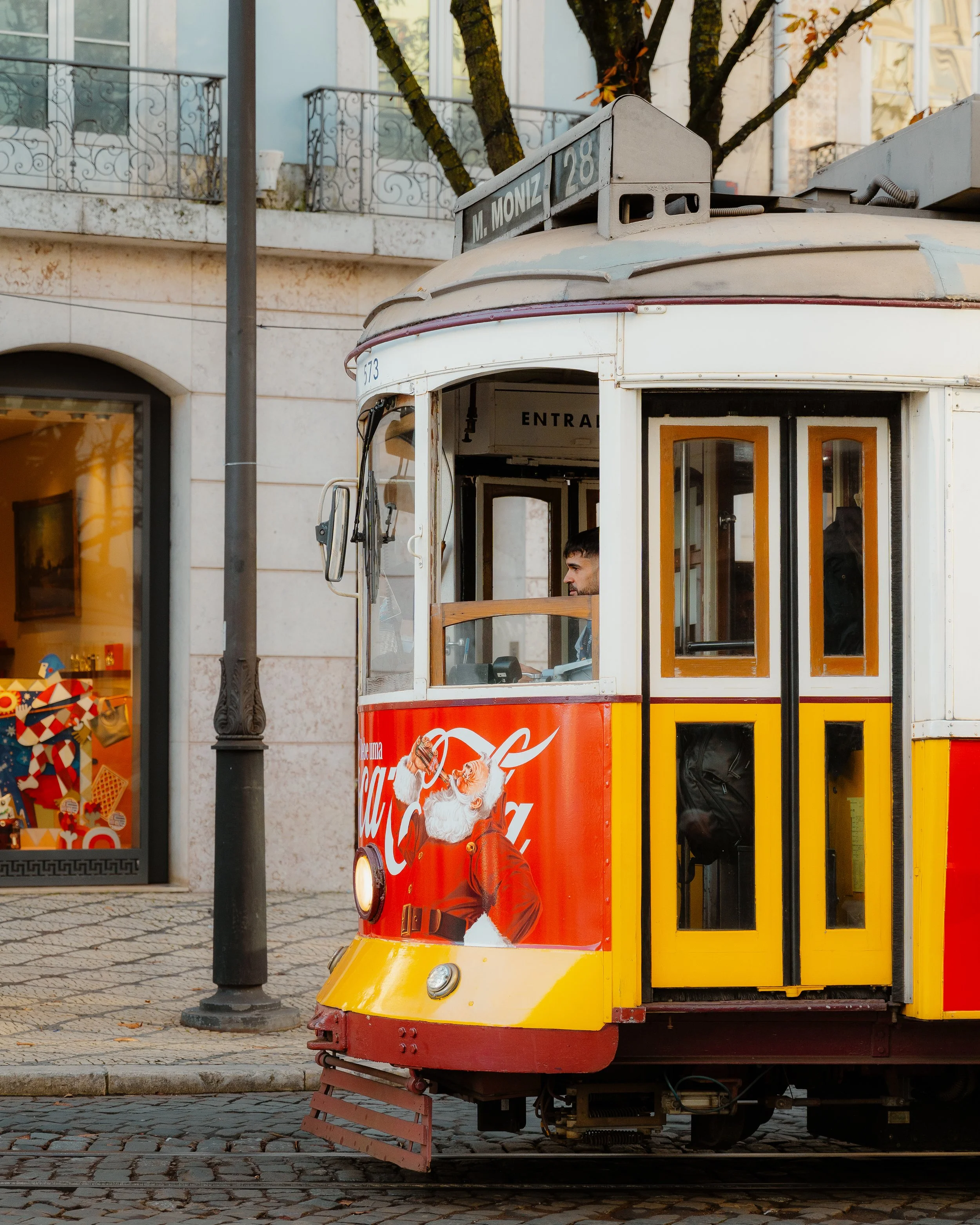 A vintage tram decorated with a Coca-Cola advertisement featuring Santa Claus, on a city street with a building and a shop window in the background.