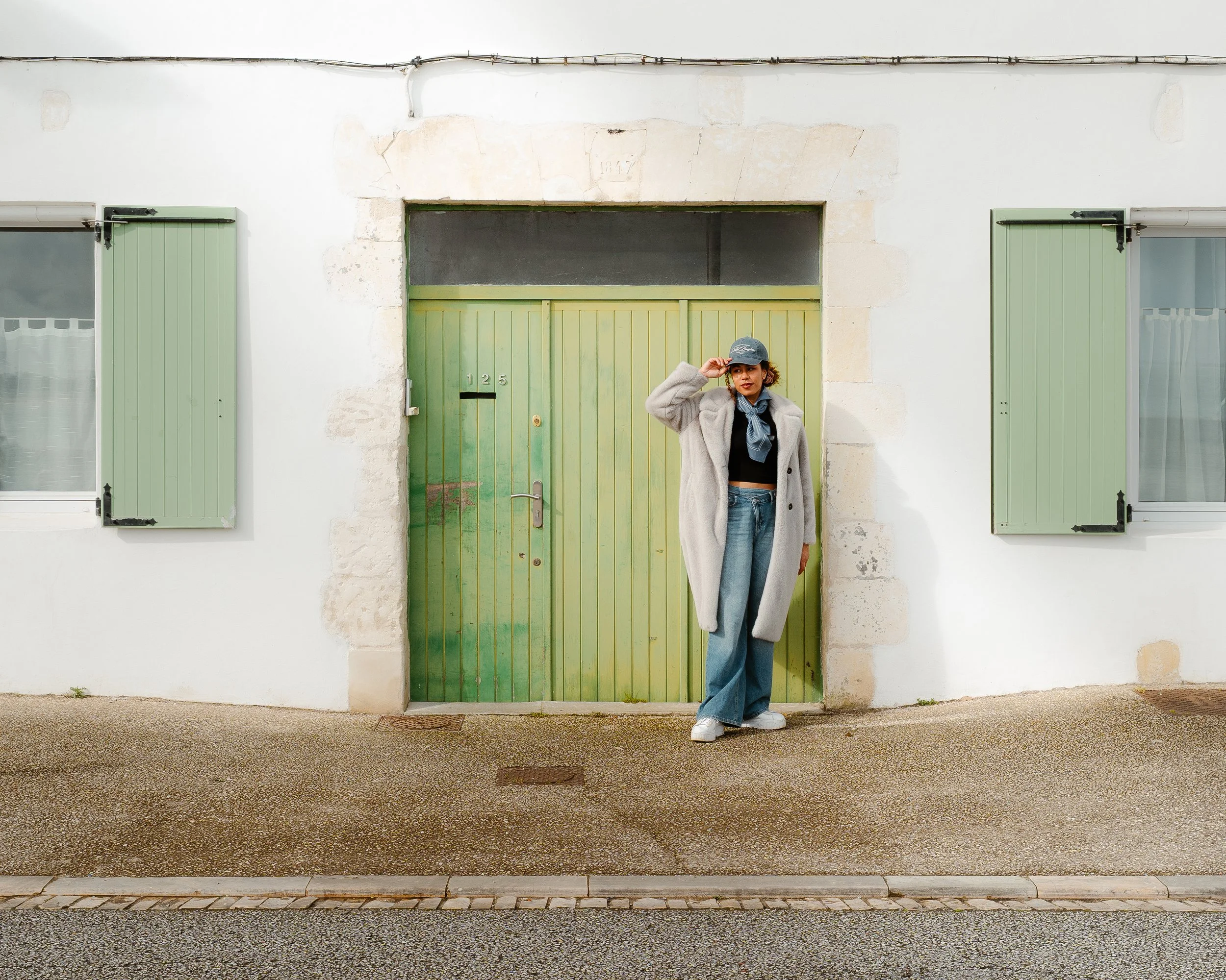 A woman standing in front of a green door with two windows on white walls, wearing a gray coat, jeans, a cap, and a scarf.