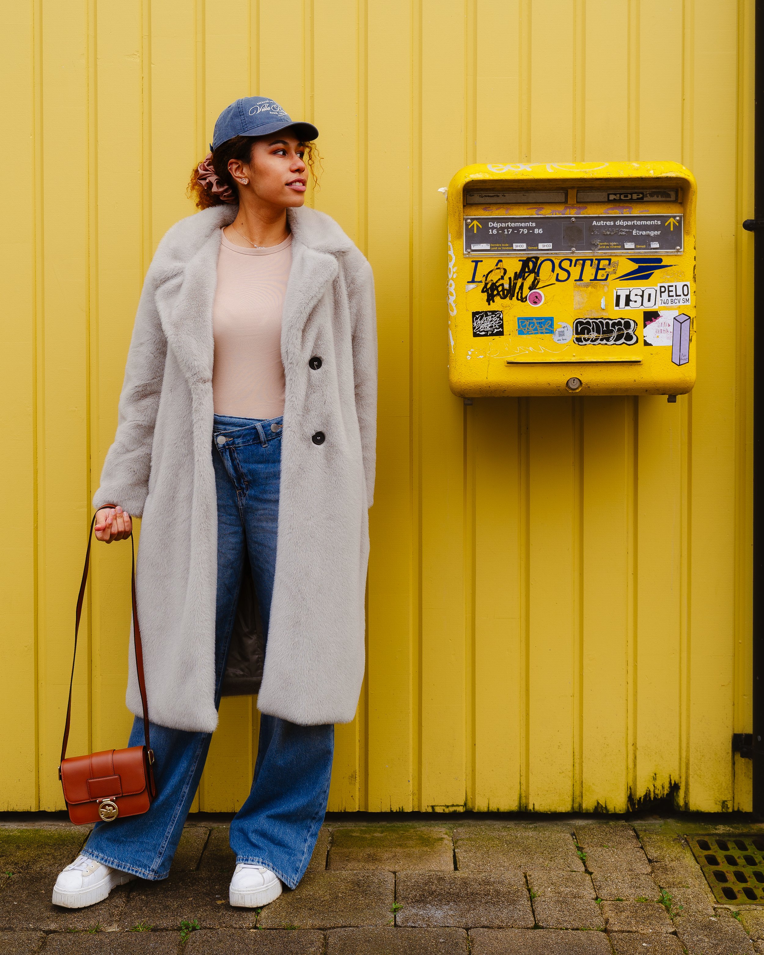 A woman standing on a sidewalk in front of a yellow wall and a yellow mailbox, wearing a beige coat, jeans, sneakers, a blue cap, and carrying a small brown purse.