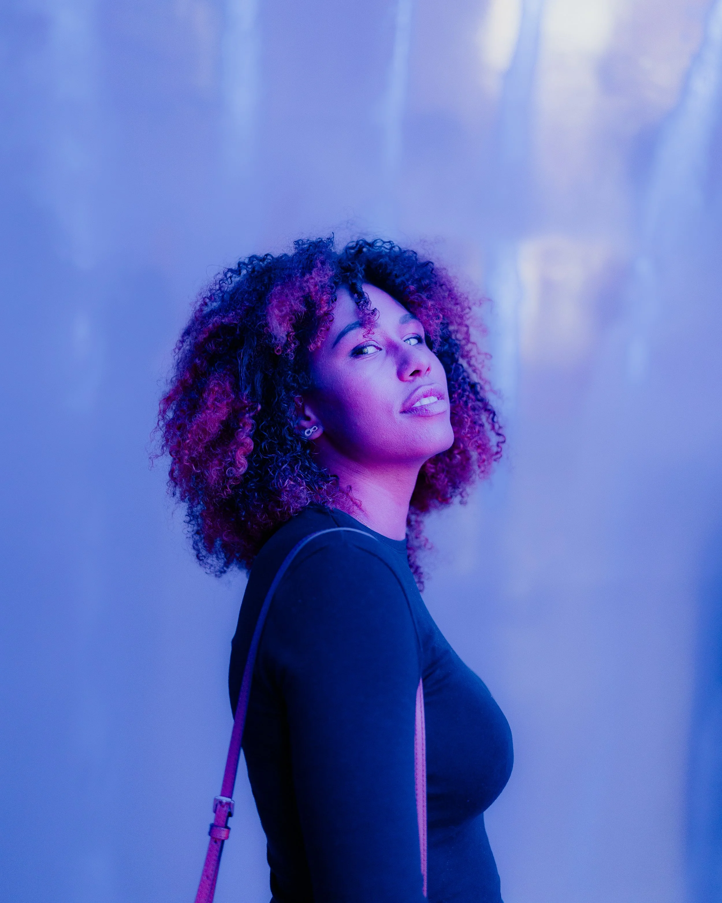 A woman with curly hair with purple and pink highlights, wearing a black shirt and earrings, standing against a blue illuminated background.