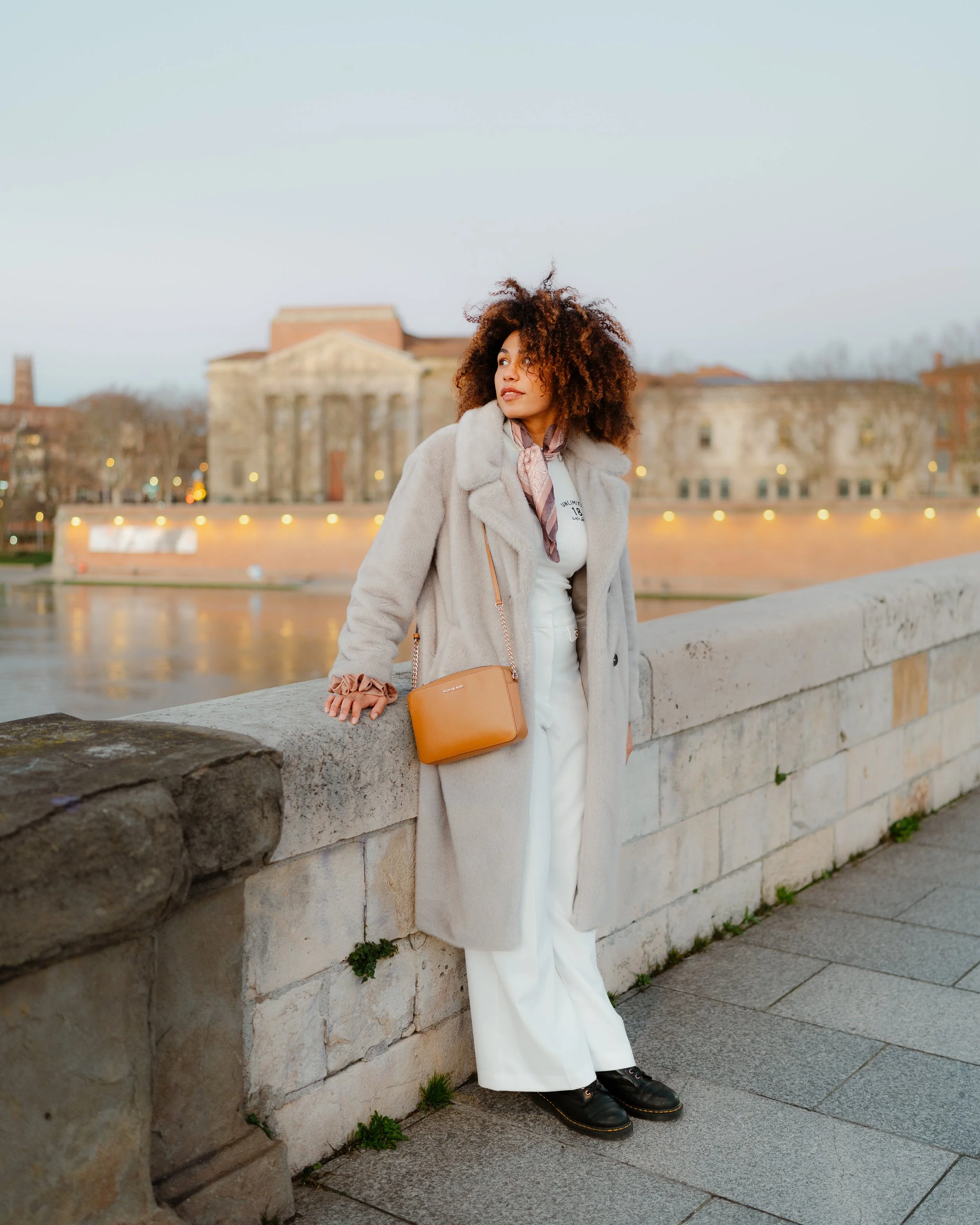 Woman with curly hair wearing a long beige coat, white pants, black shoes, and a patterned scarf, standing beside a stone wall with a river and classical building in the background during evening.