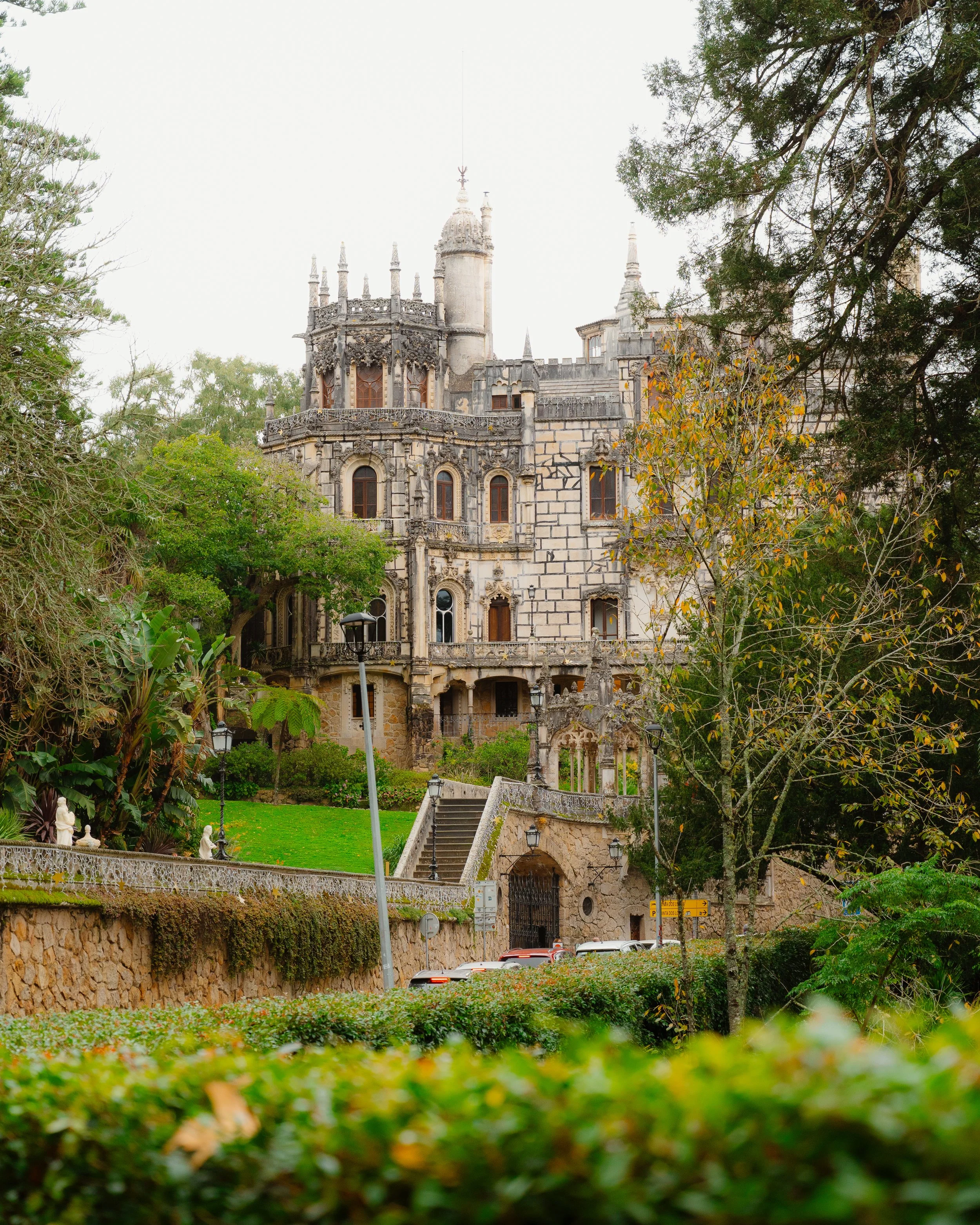 A historic castle with gothic architecture, surrounded by trees and greenery, with a staircase leading up to the entrance.