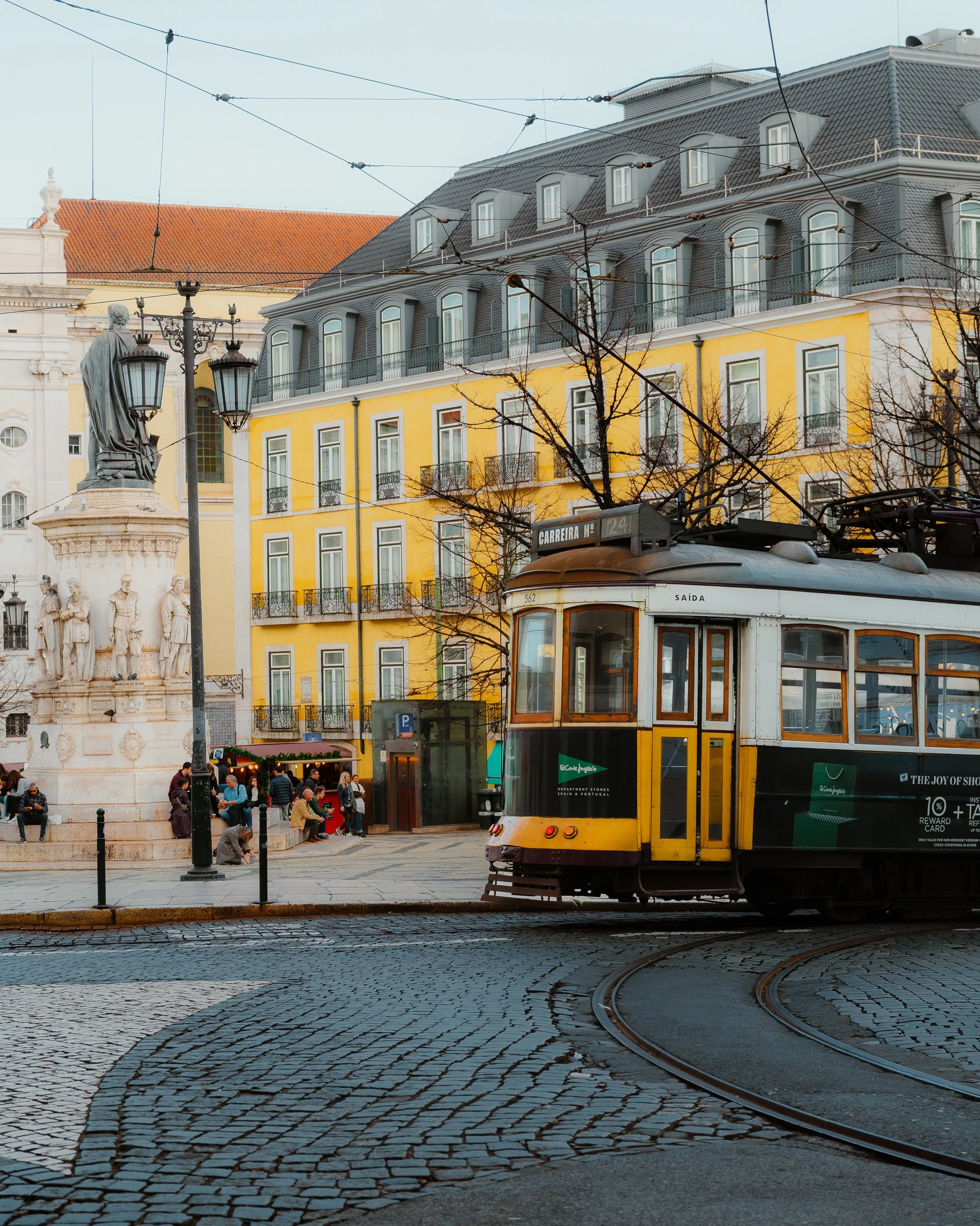 A street scene with a yellow and gray building, a tram in the foreground, and a statue with a fountain and people near it.