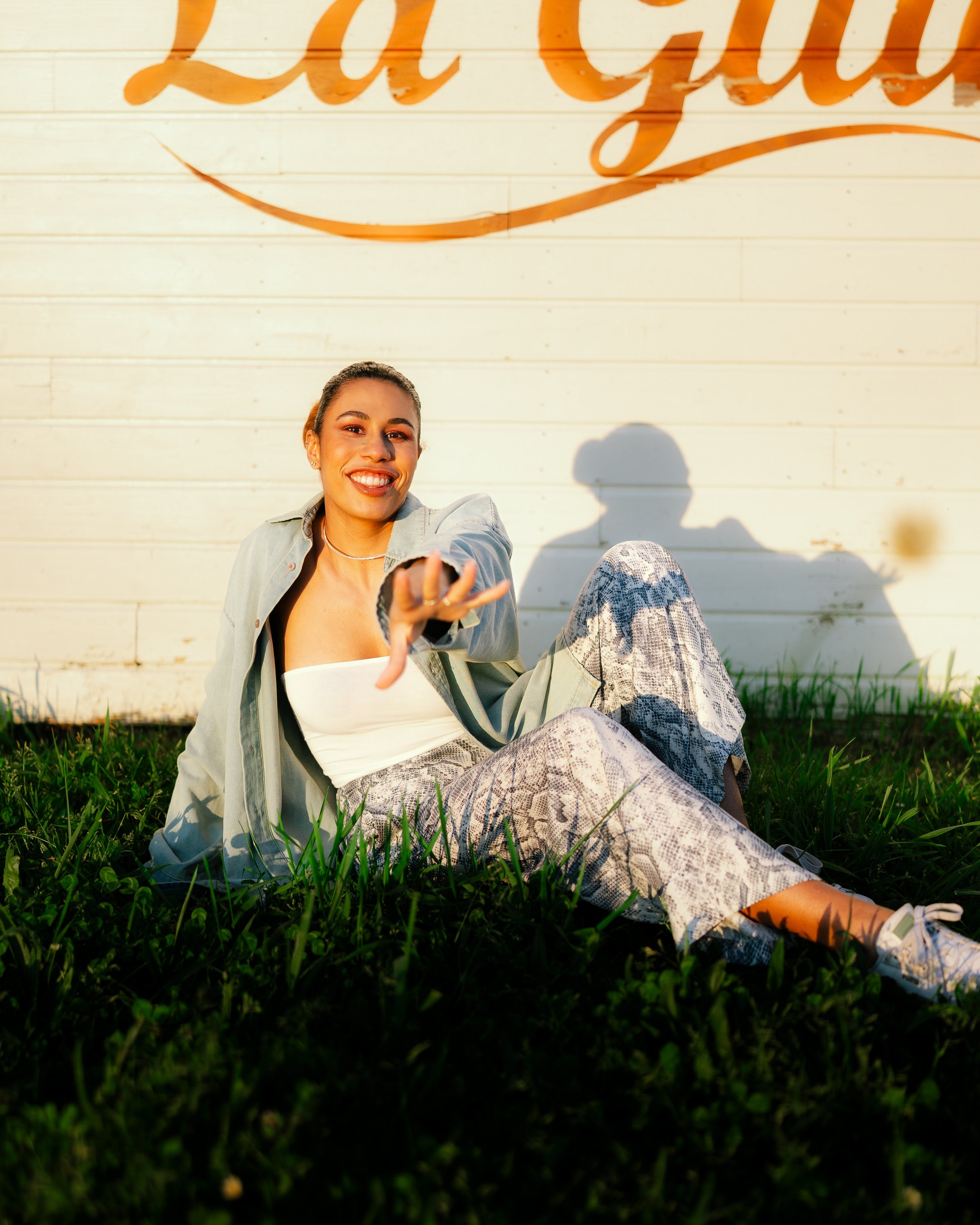 Young woman sitting on grass in front of a white wooden wall with orange lettering, smiling, reaching out, with shadows cast on the wall.