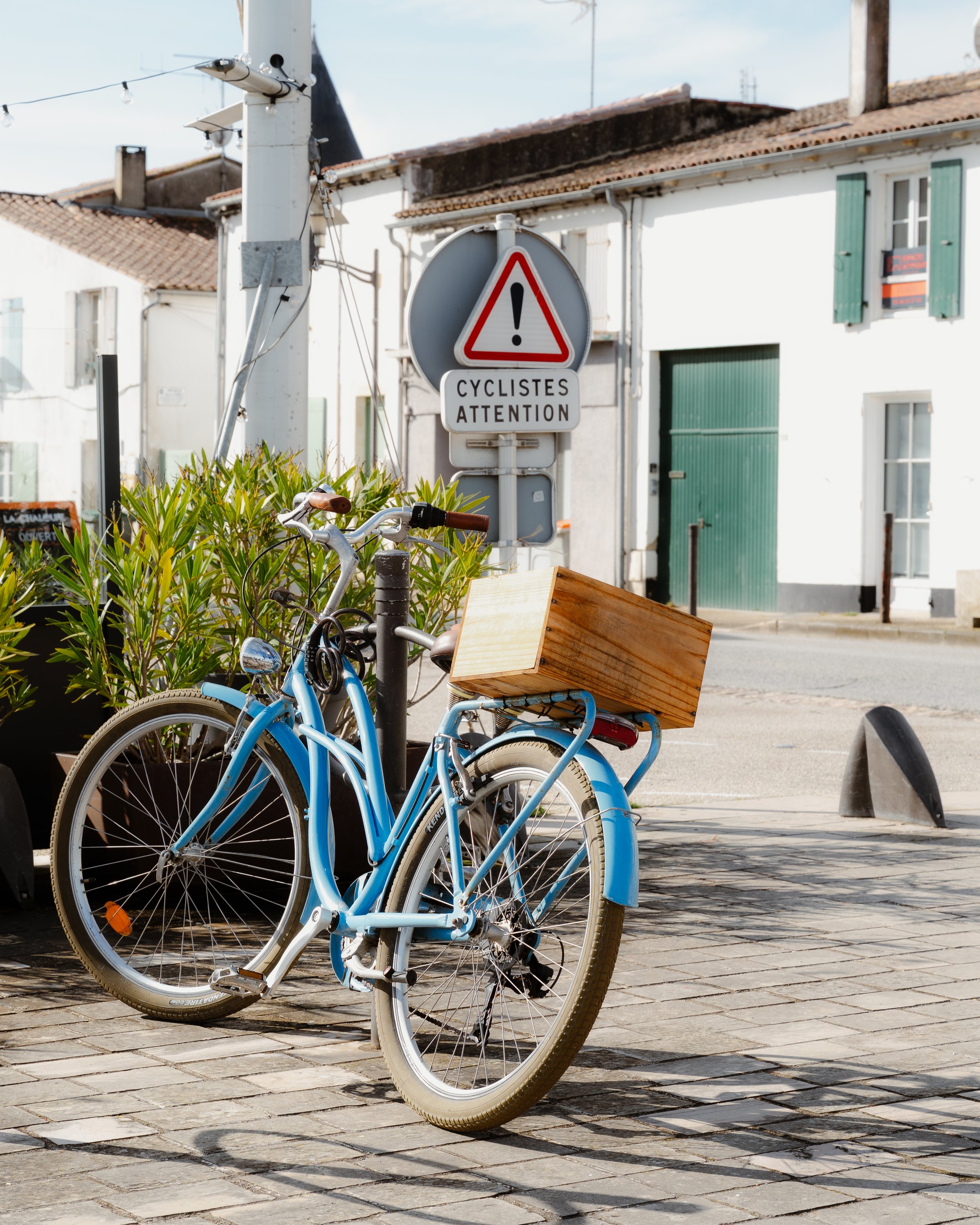 A blue bicycle with a wooden box attached to the back, parked beside a street with plants and a traffic sign that reads 'Cyclistes Attention' and an exclamation mark.