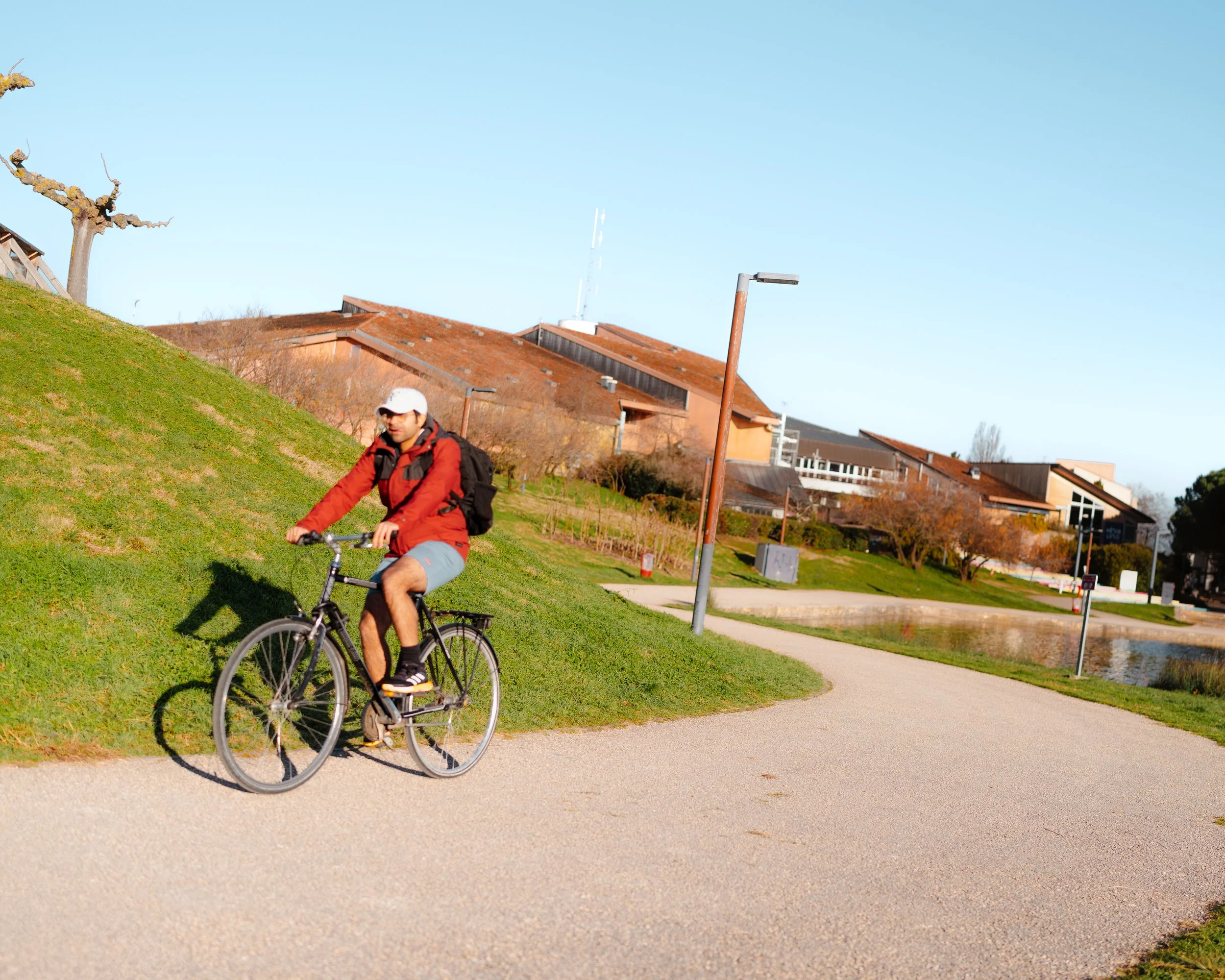 A man riding a bicycle on a paved path beside a grassy hill in a park, with houses and a pond in the background, on a sunny day.
