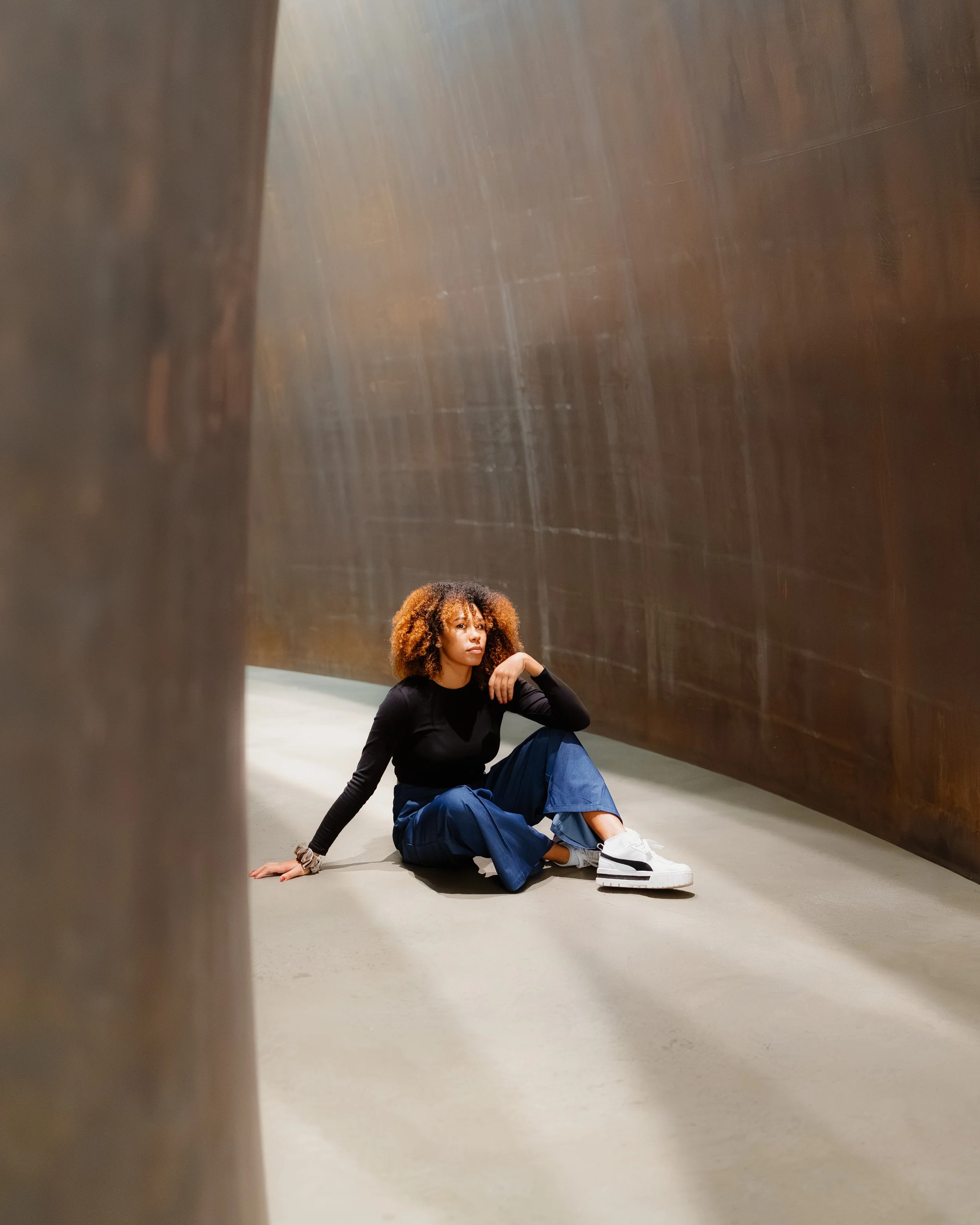Woman with curly hair sitting on the floor in a narrow passageway between two large, bronze-colored walls.