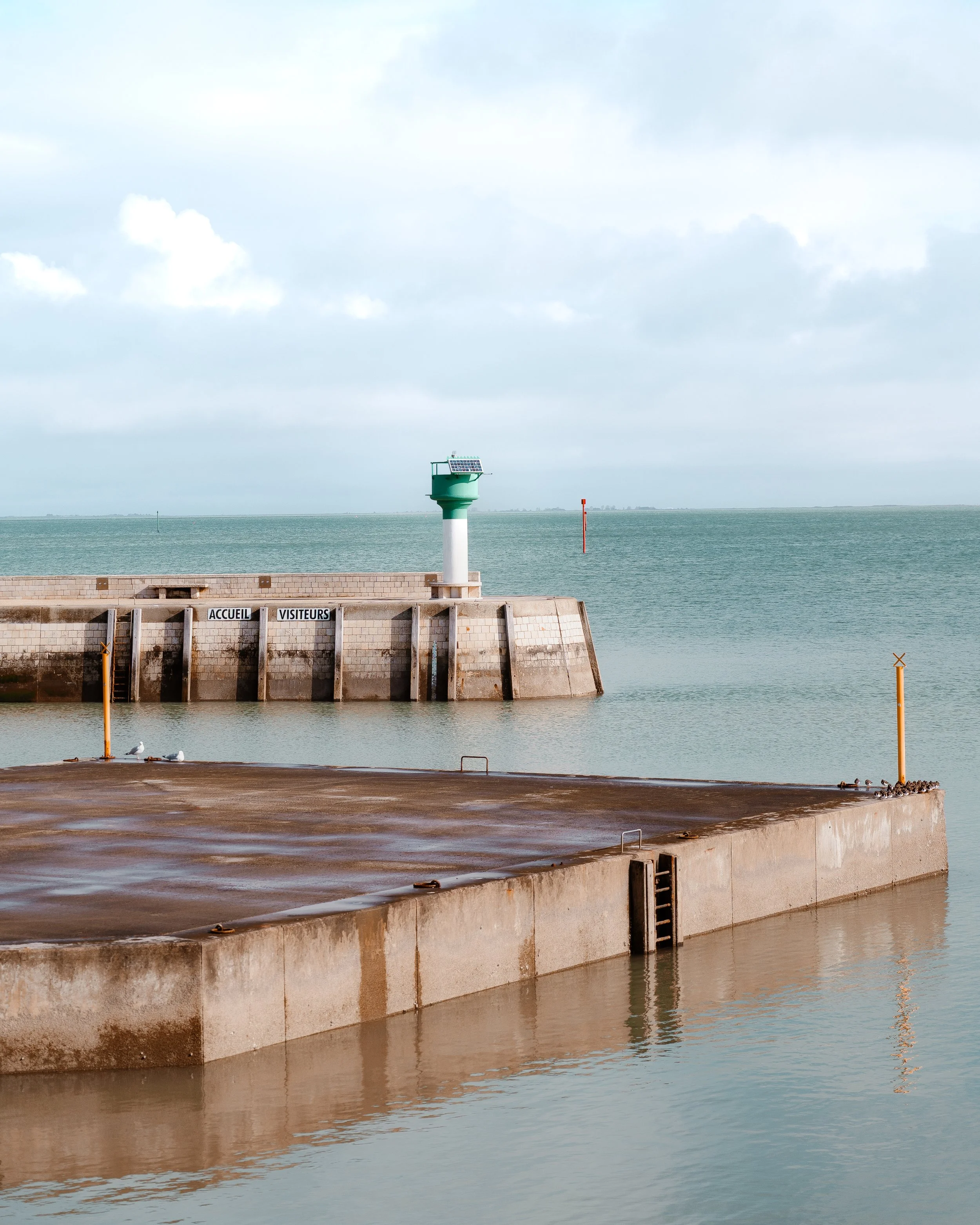 A calm harbor with a concrete pier, a small green lighthouse, and the ocean in the background under a cloudy sky.