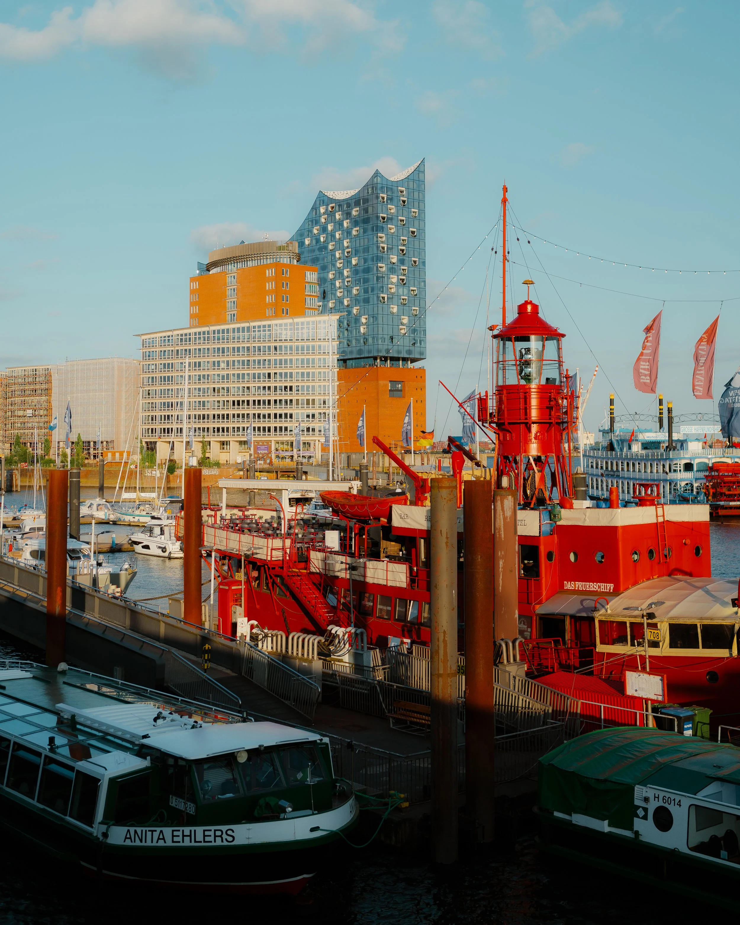 A harbor scene with boats docked, including a bright red ship with a lighthouse structure, and modern buildings in the background under a partly cloudy sky.