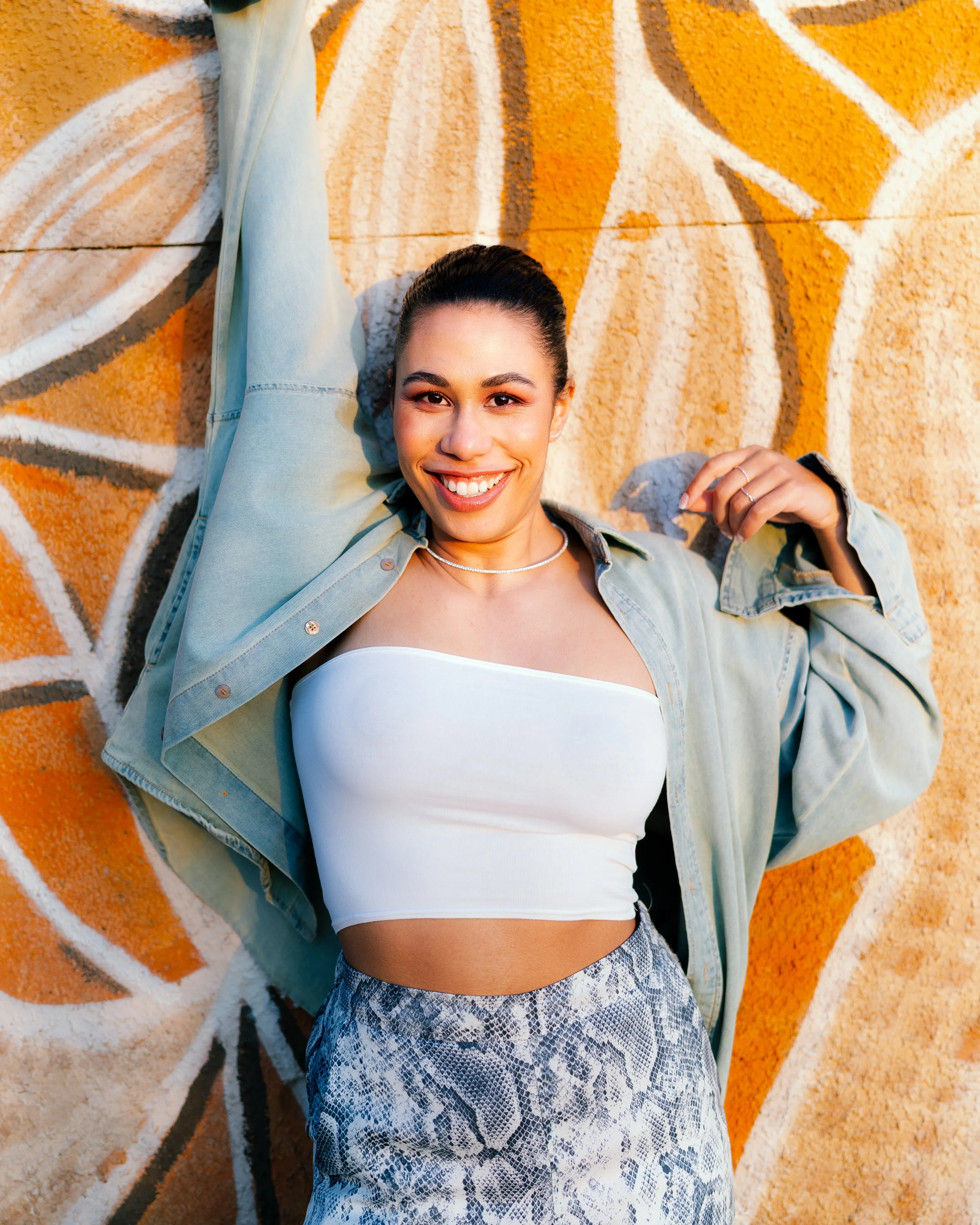 A smiling woman with dark hair tied back, wearing a white tube top, a light green jacket, and snakeskin-patterned pants, lying against a graffiti-covered wall with orange and white designs.