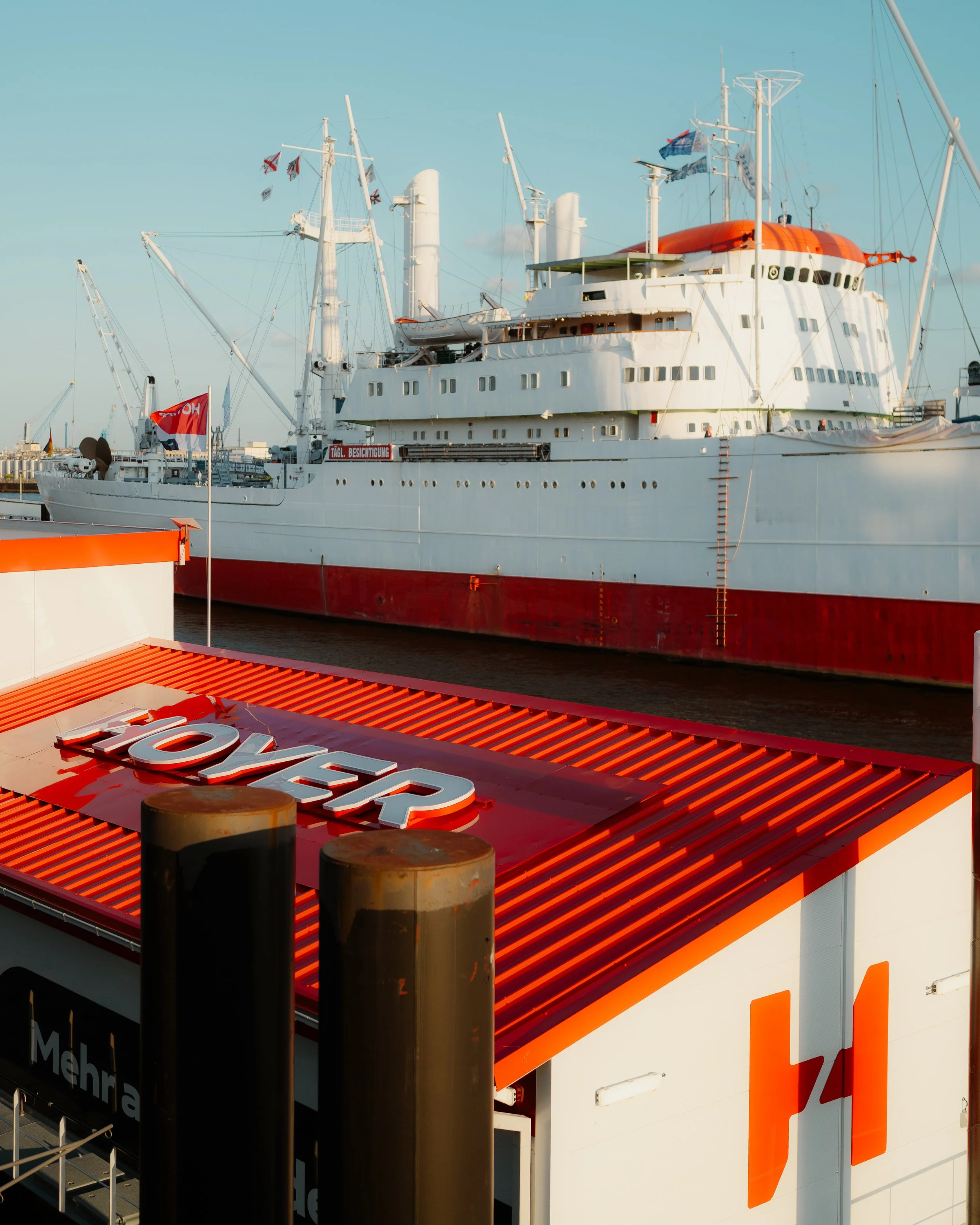 A large white cruise ship docked at a harbor with a red hull and orange accents. In the foreground, there is a sign with the word 'LOVER' on a red and white structure.