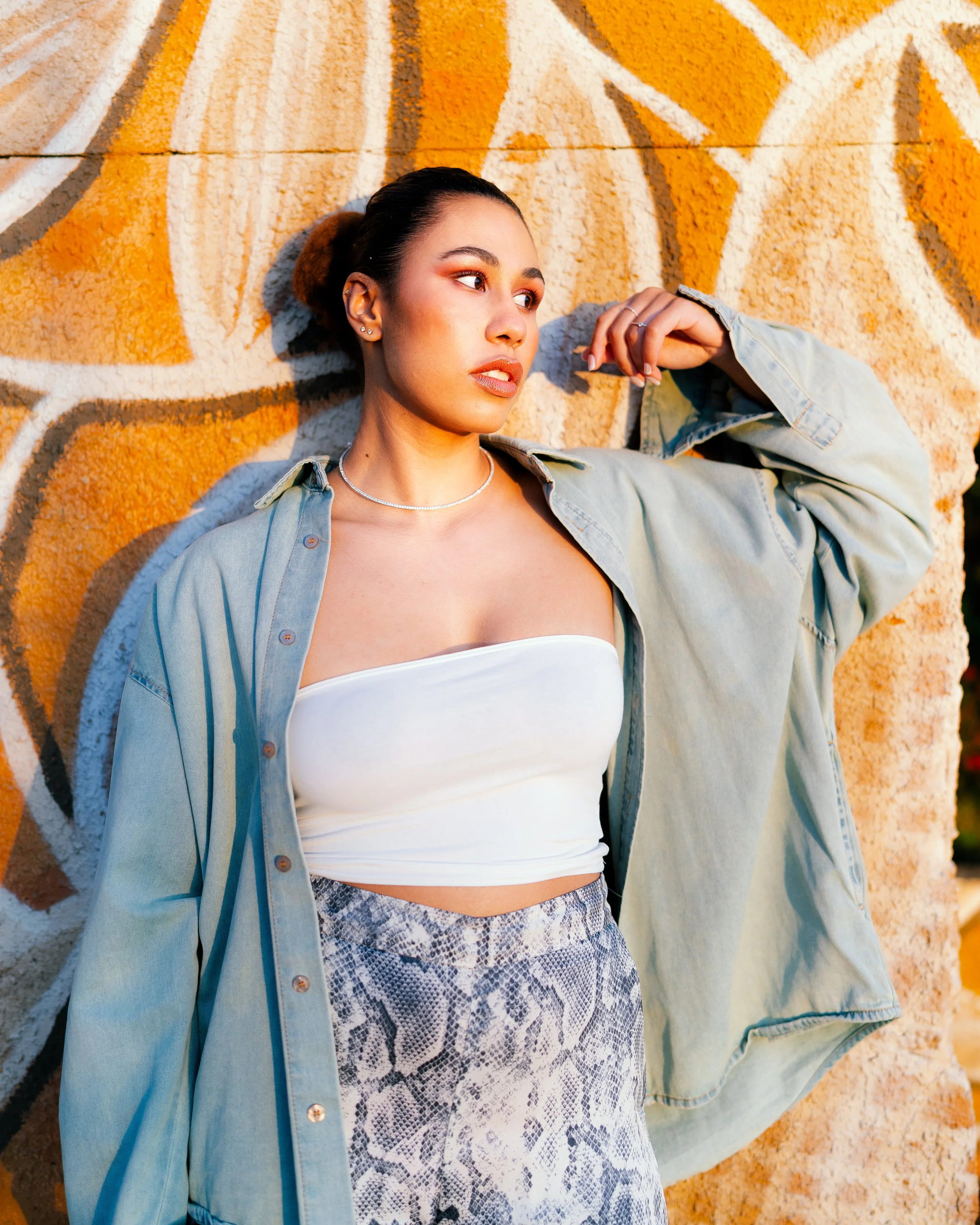 A young woman with dark hair in a bun, wearing a white tube top, patterned pants, and an oversized denim jacket, stands in front of a colorful graffiti wall.