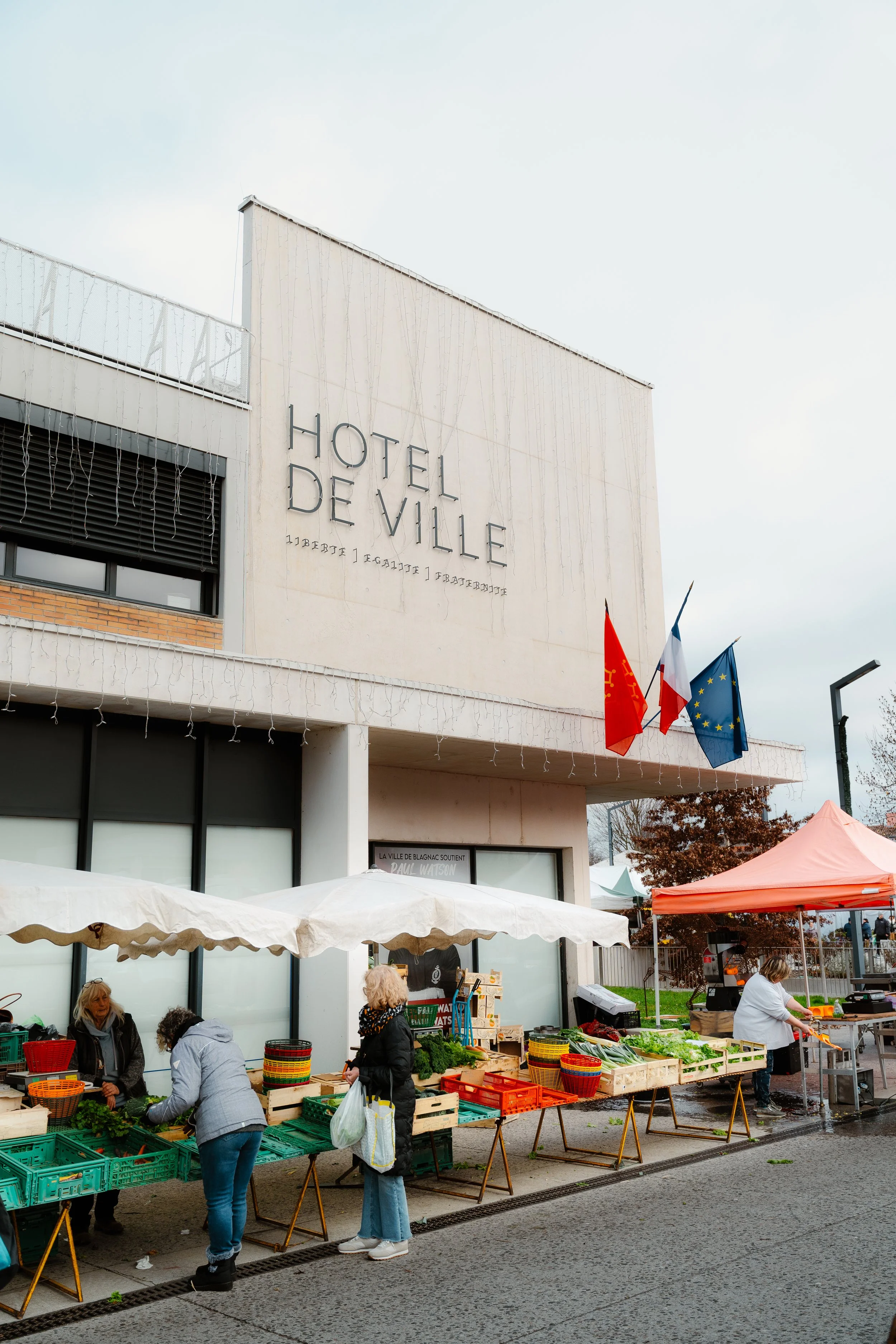 An outdoor vegetable market in front of Hotel de Ville with people buying produce.