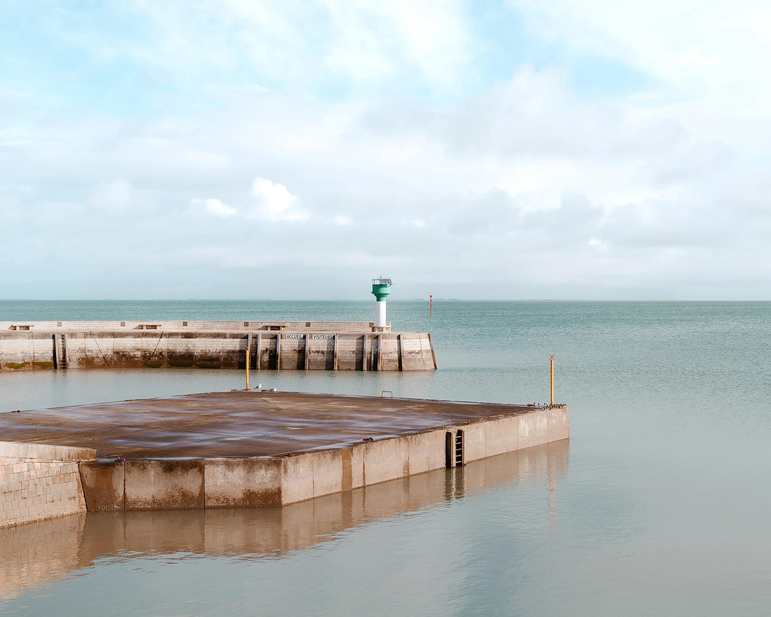 A harbor with concrete piers extending into the calm sea, a green and white lighthouse on one pier, and a cloudy sky overhead.